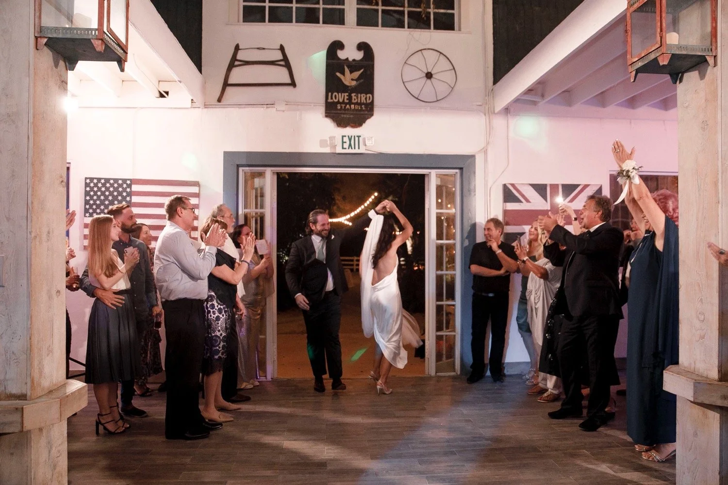 Wedding reception with bride and groom dancing, guests clapping and cheering in a rustic indoor venue decorated with American flags and vintage farm tools, nighttime outdoor scene visible through open doors.