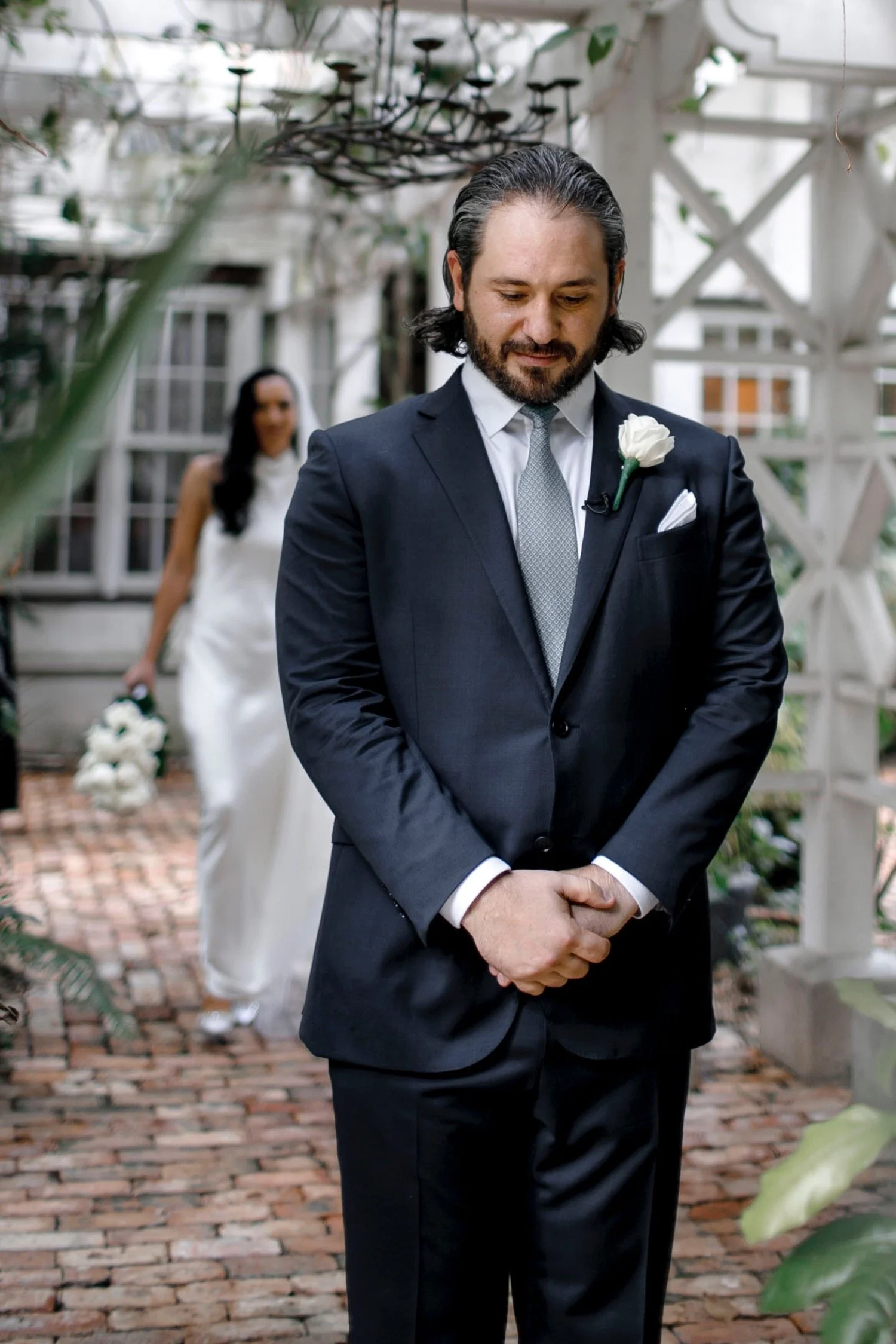 A groom in a dark suit with a white boutonniere and a gray tie stands with his hands clasped in front of him, looking down, during a wedding ceremony. A bride in a white dress holding a bouquet of white flowers is approaching in the background in a g