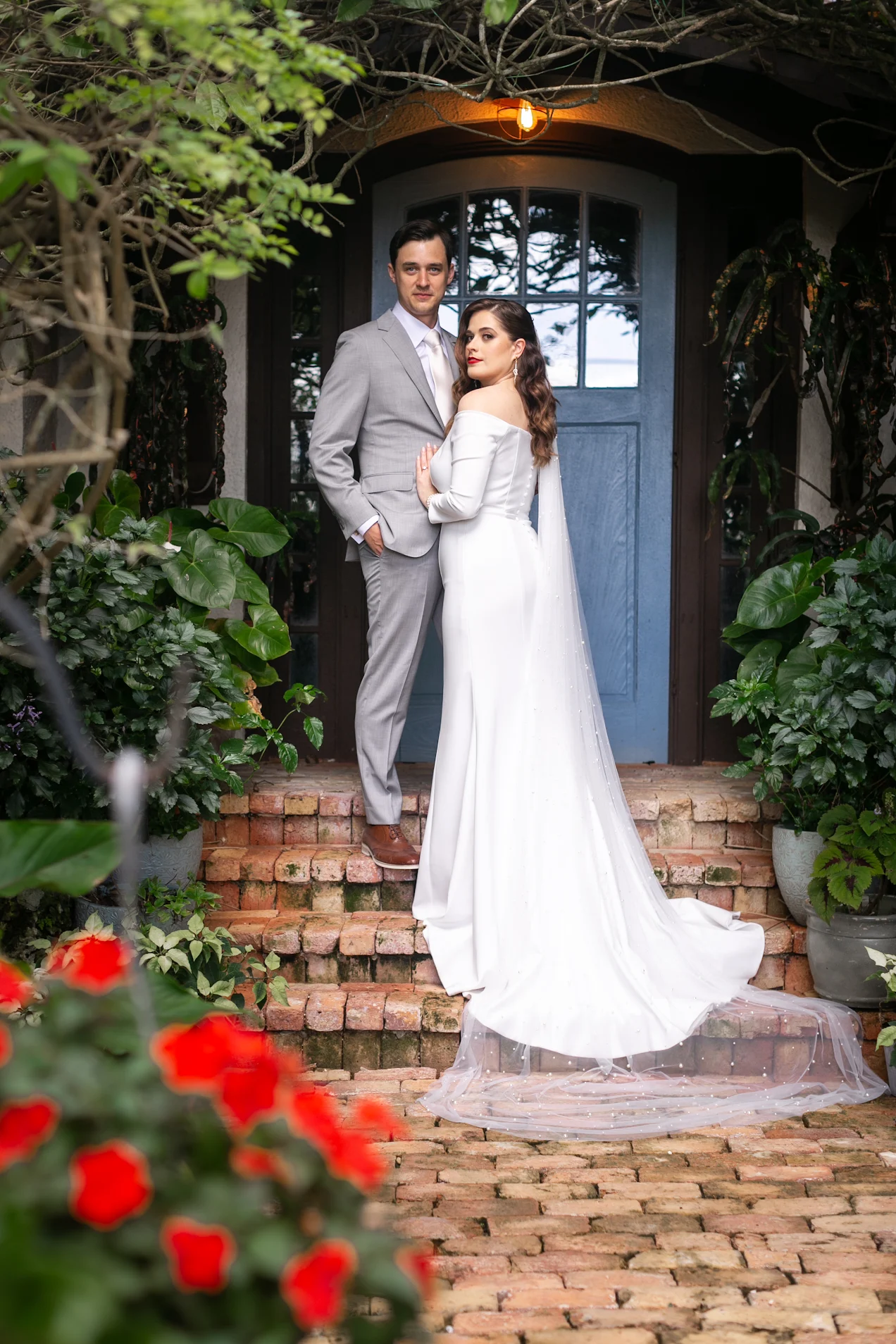 A bride in a white wedding gown and a groom in a gray suit stand together on brick stairs outside a house with a blue door, surrounded by lush green plants and red flowers in the foreground.