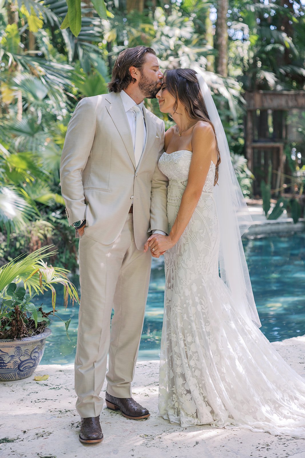 A bride and groom holding hands by a pool in a lush tropical garden, with the groom kissing the bride's forehead during their wedding.