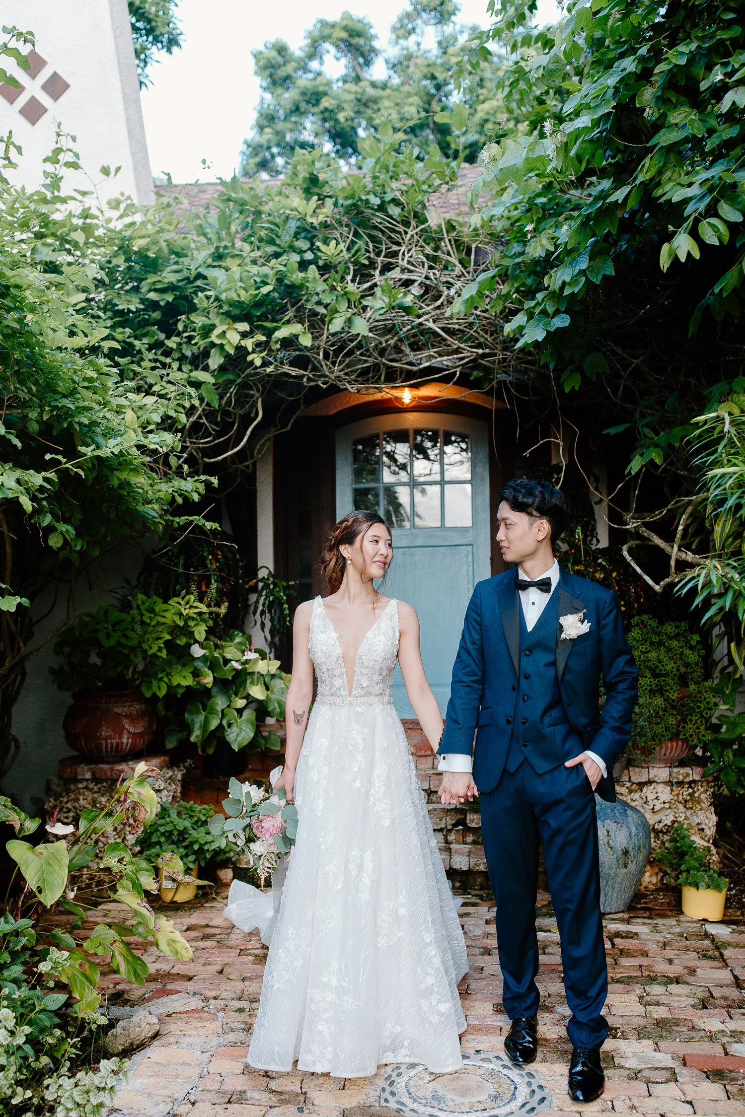 Bride and groom holding hands, standing outdoors on brick pathway, surrounded by lush greenery, in front of a rustic door with window, wedding attire, romantic atmosphere.