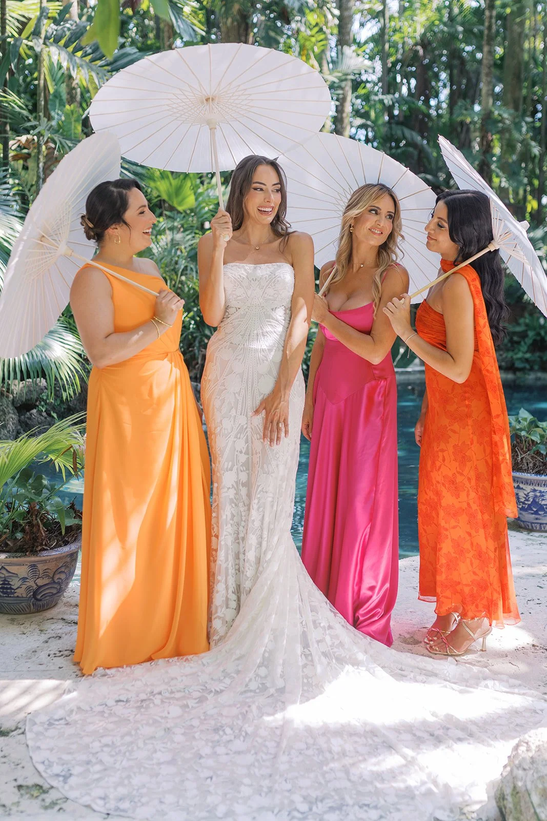 Four women in colorful dresses holding white parasols are standing outdoors with tropical plants and a pool in the background. One woman wears a white lace wedding dress, and the other three wear vibrant pink and orange dresses, smiling and chatting.