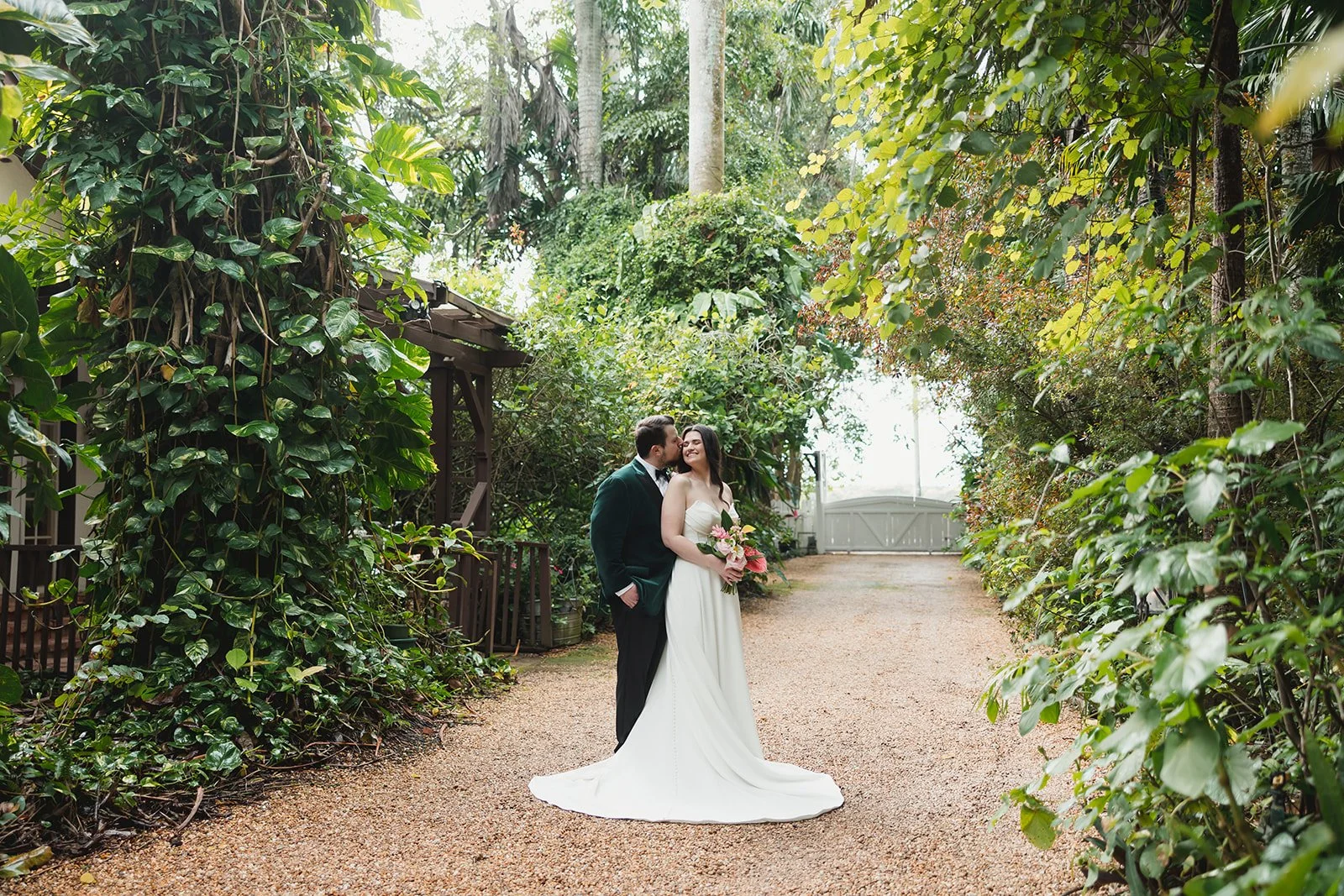 A bride and groom standing close together in a lush, green outdoor setting, with the groom kissing the bride on the cheek while she holds a bouquet of pink and white flowers. The bride is wearing a white wedding dress and the groom a dark suit. They 