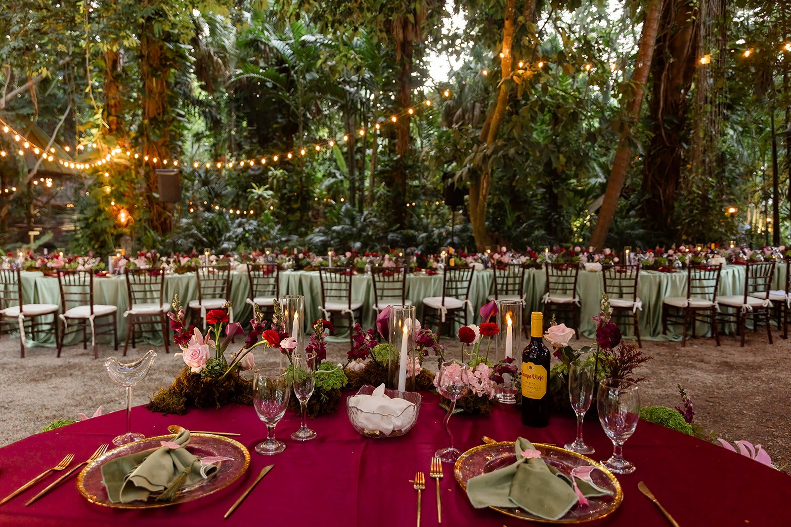 A beautifully decorated outdoor event space with a long banquet table and chairs, surrounded by lush greenery. String lights hang above, creating a warm ambiance. The table features candles, flowers, wine bottles, and neatly arranged glassware and cu
