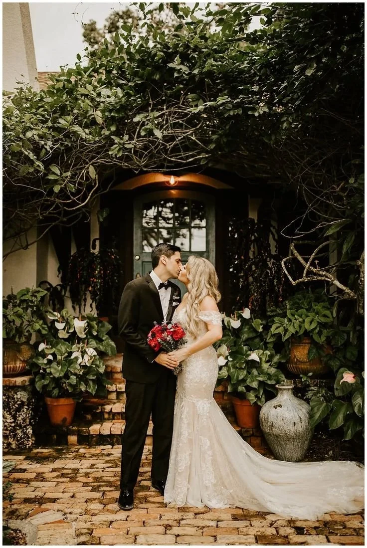 A bride and groom share a kiss outdoors. The bride wears a white lace wedding gown and holds a bouquet of red roses. The groom is dressed in a black tuxedo with a bow tie. They stand on a brick path in front of a dark door surrounded by lush green pl