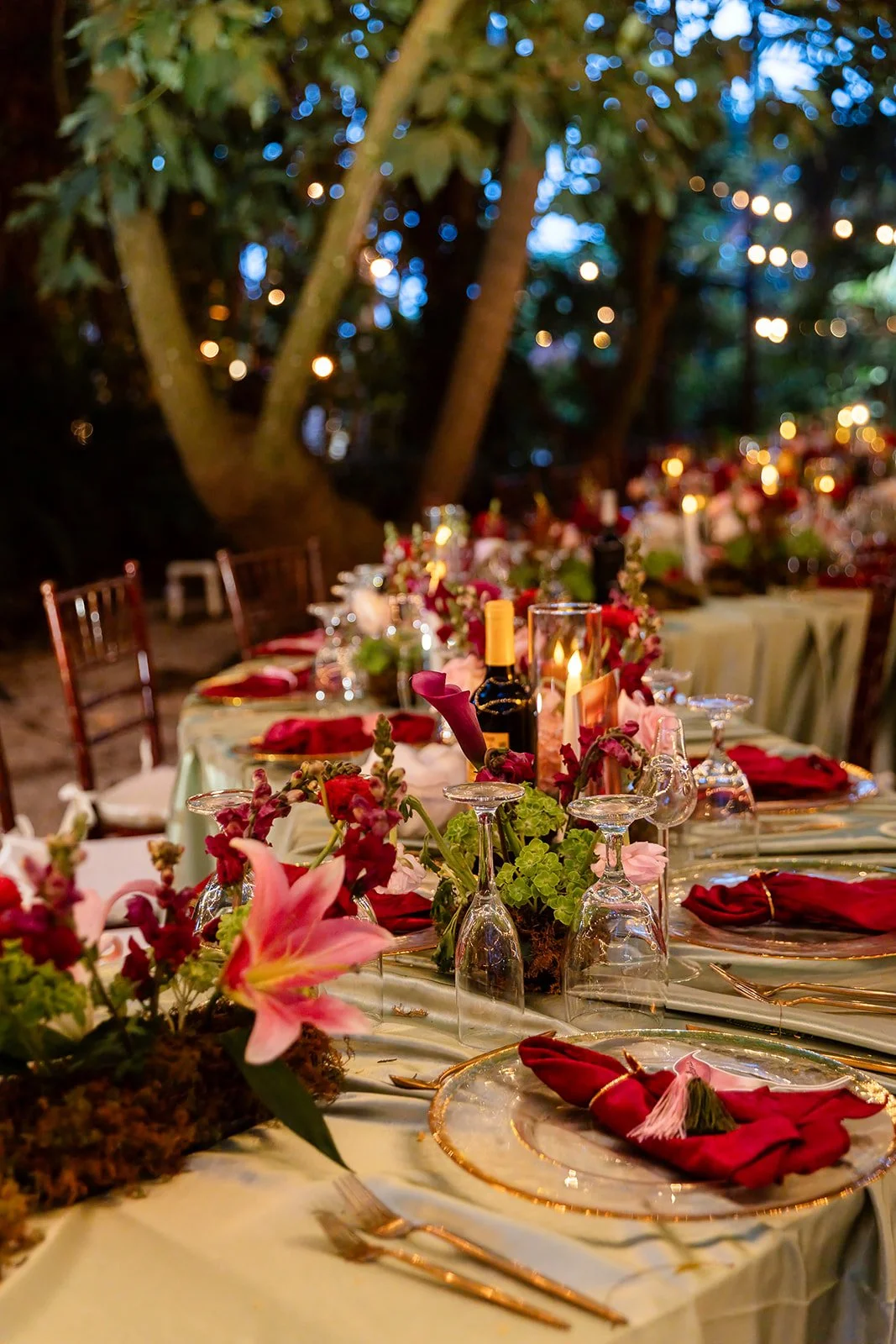 An elegant outdoor dinner table decorated with floral arrangements, candles, wine, and tableware, set amidst trees with blurred string lights in the background during evening.