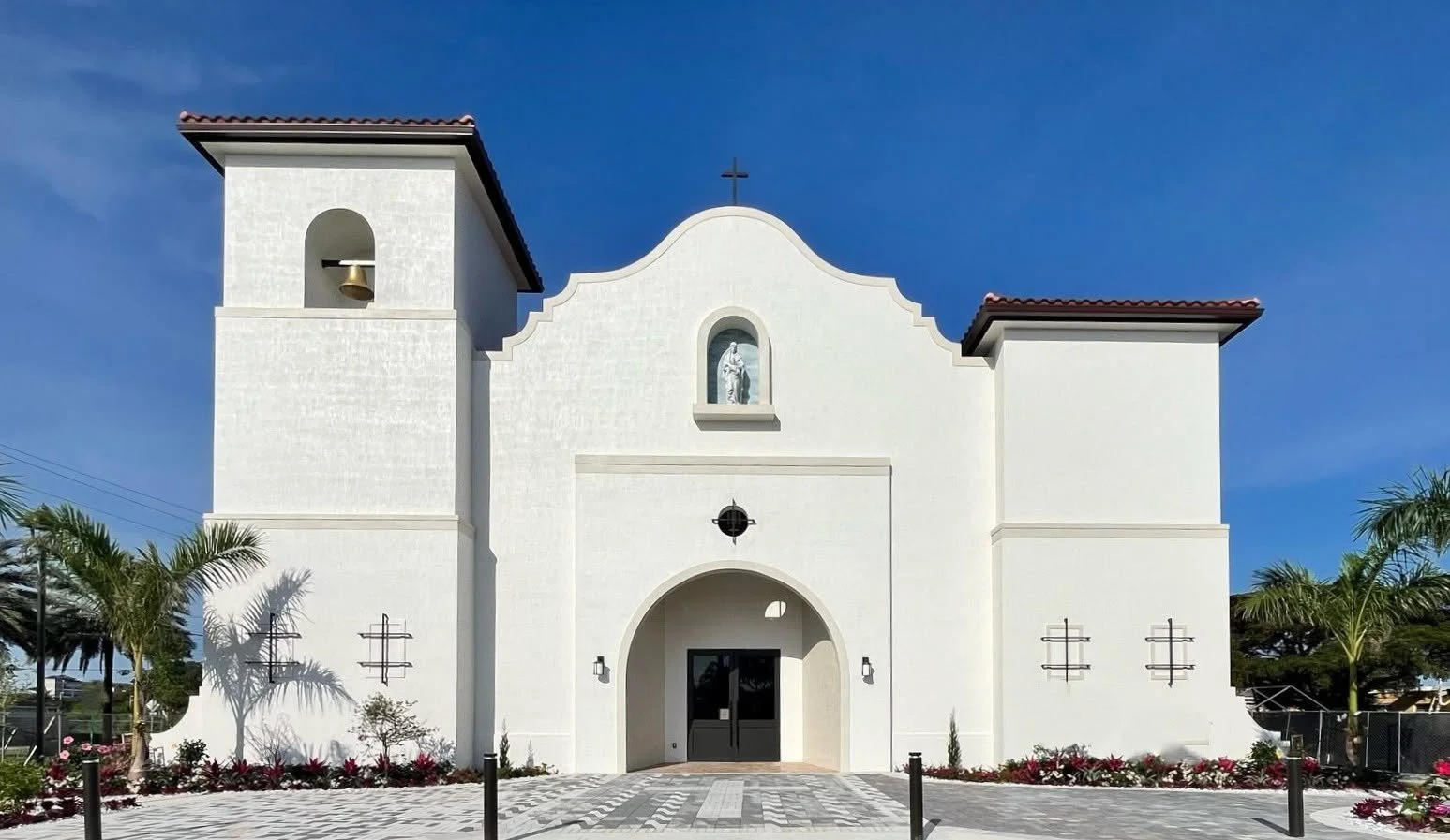 Front view of a white church with a bell tower on the left, a statue in a niche above the main entrance, and a cross on top of the curved roof. The church is surrounded by palm trees and colorful flowers, under a clear blue sky.