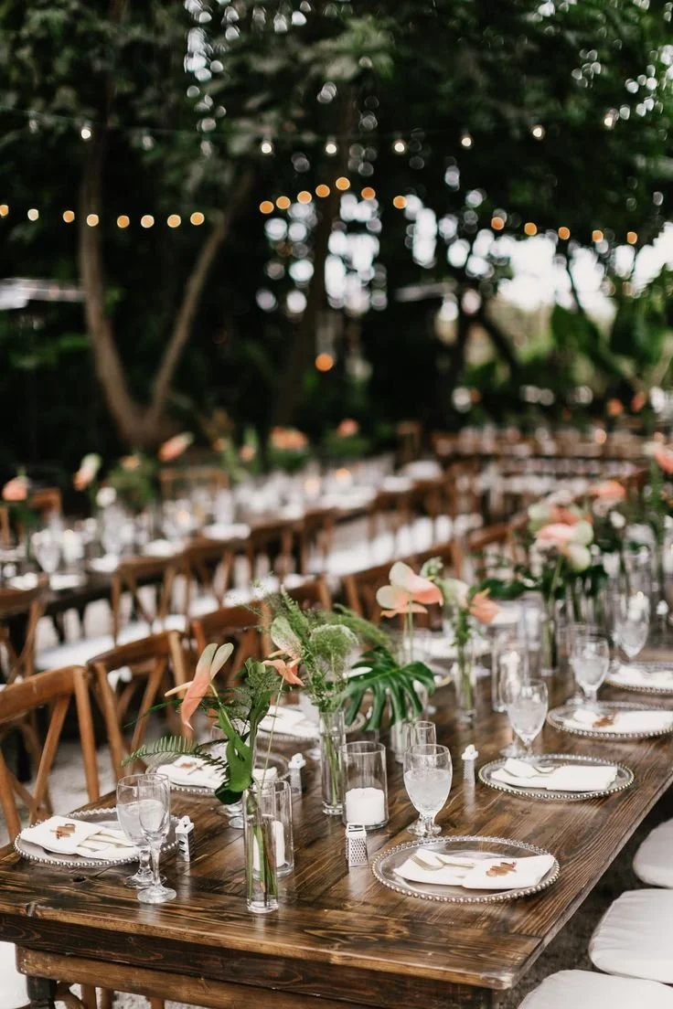 Outdoor dining table decorated with floral centerpieces, glassware, and elegant table settings, surrounded by trees and string lights for an event or celebration.