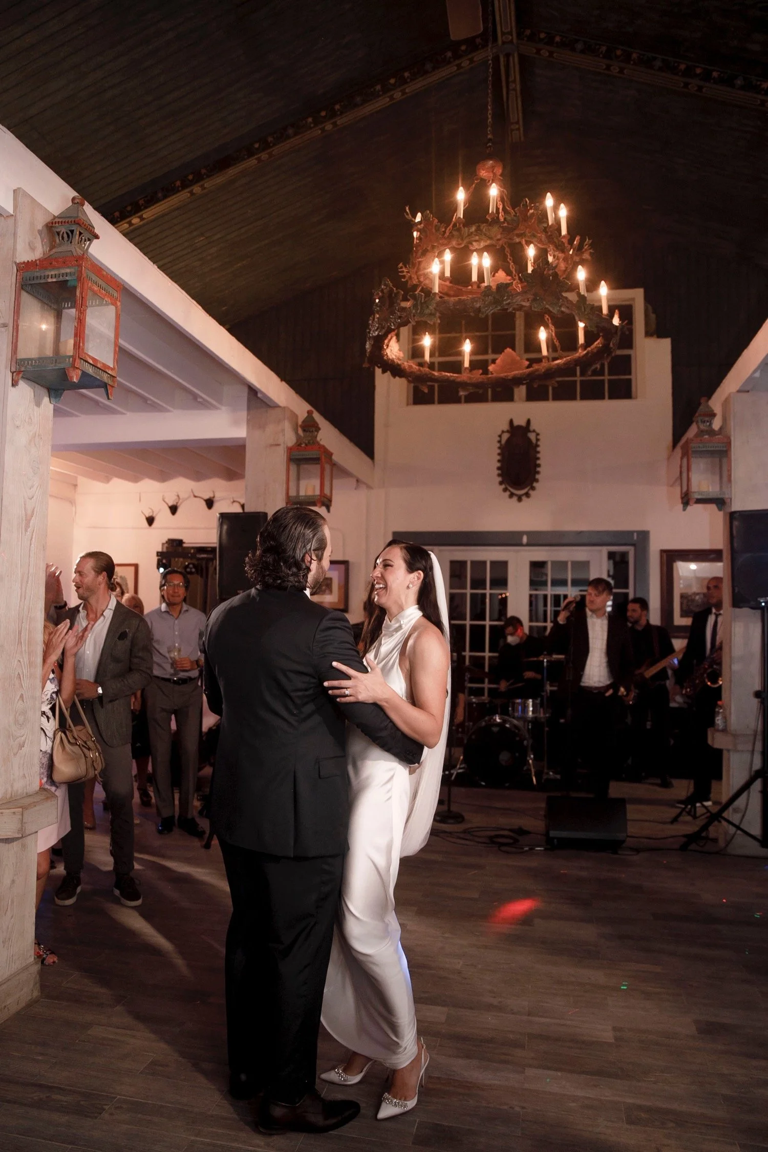 Bride and groom dancing at a wedding reception with guests and a live band in the background, under a rustic chandelier.