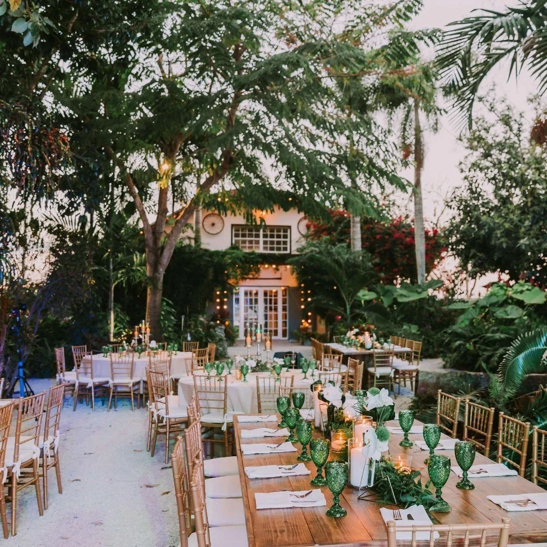 Outdoor event setup with round and rectangular tables, decorated with green glassware, white napkins, and candles, surrounded by wooden chairs on a sandy ground amid tropical trees and plants, with a house-like structure in the background.