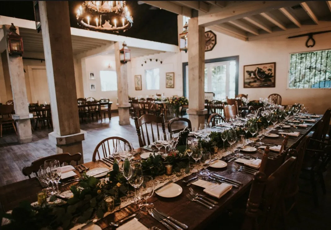 A decorated dining area with a long table set for a special occasion, featuring wine glasses, plates, silverware, and greenery centerpieces, inside a rustic-style room with wooden beams, framed artwork, and large windows.