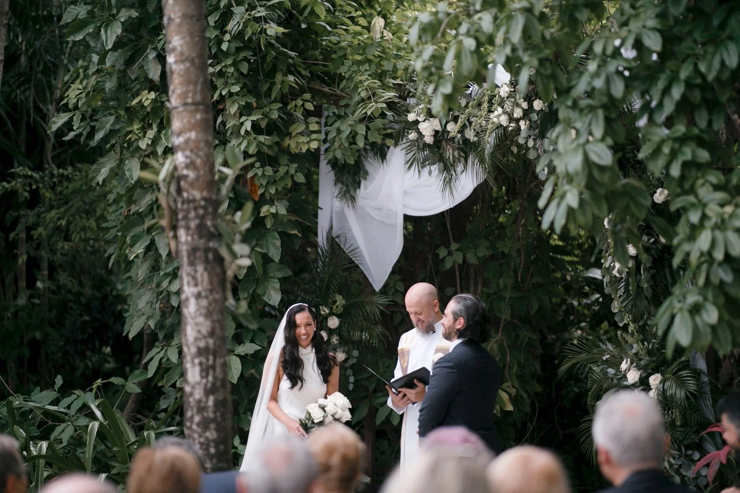 A wedding ceremony taking place outdoors in a lush, green garden with a bride holding a bouquet, standing before a groom and officiant, surrounded by guests.