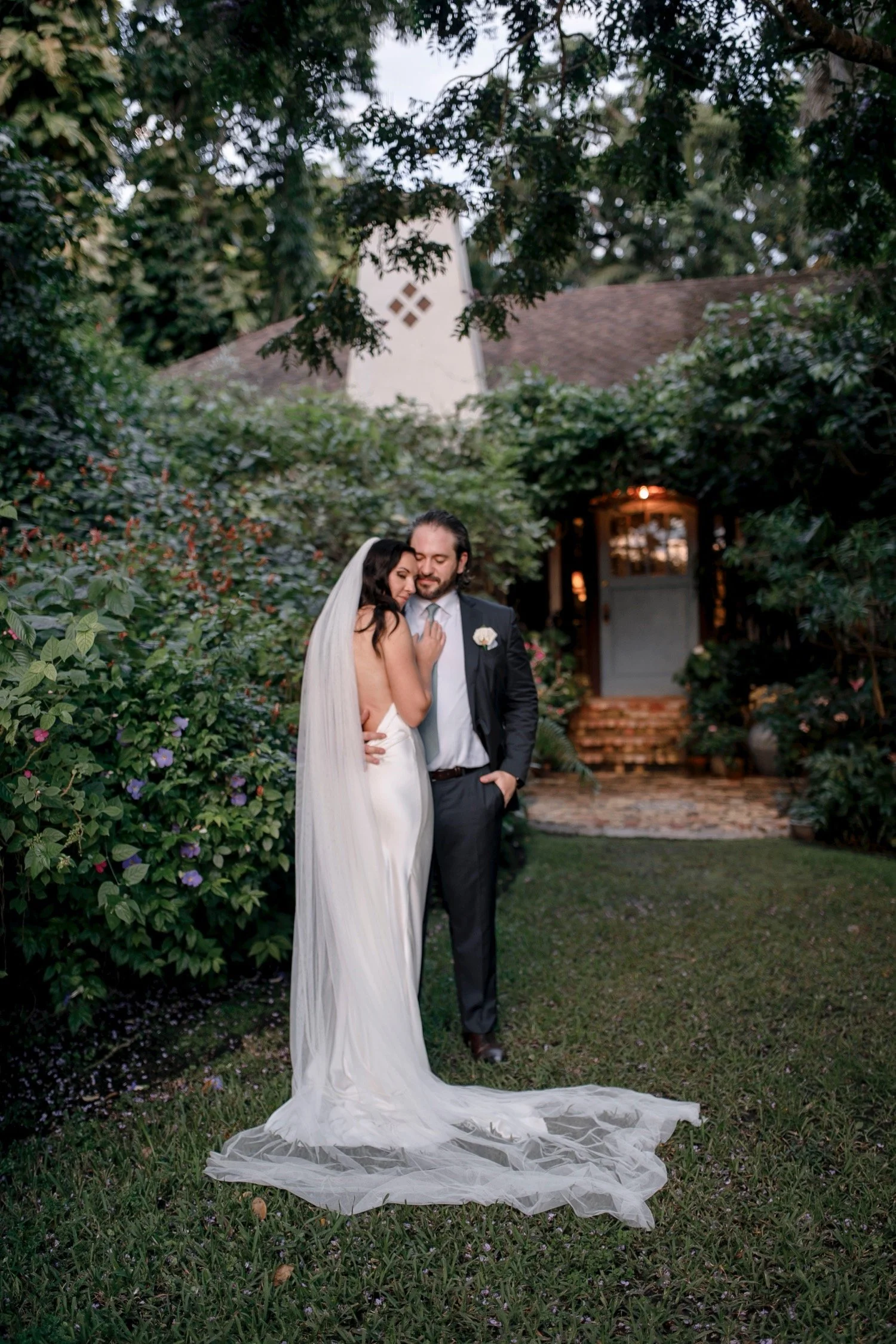 A bride and groom stand close together outdoors among lush greenery and colorful flowers, with a house in the background, during their wedding celebration.