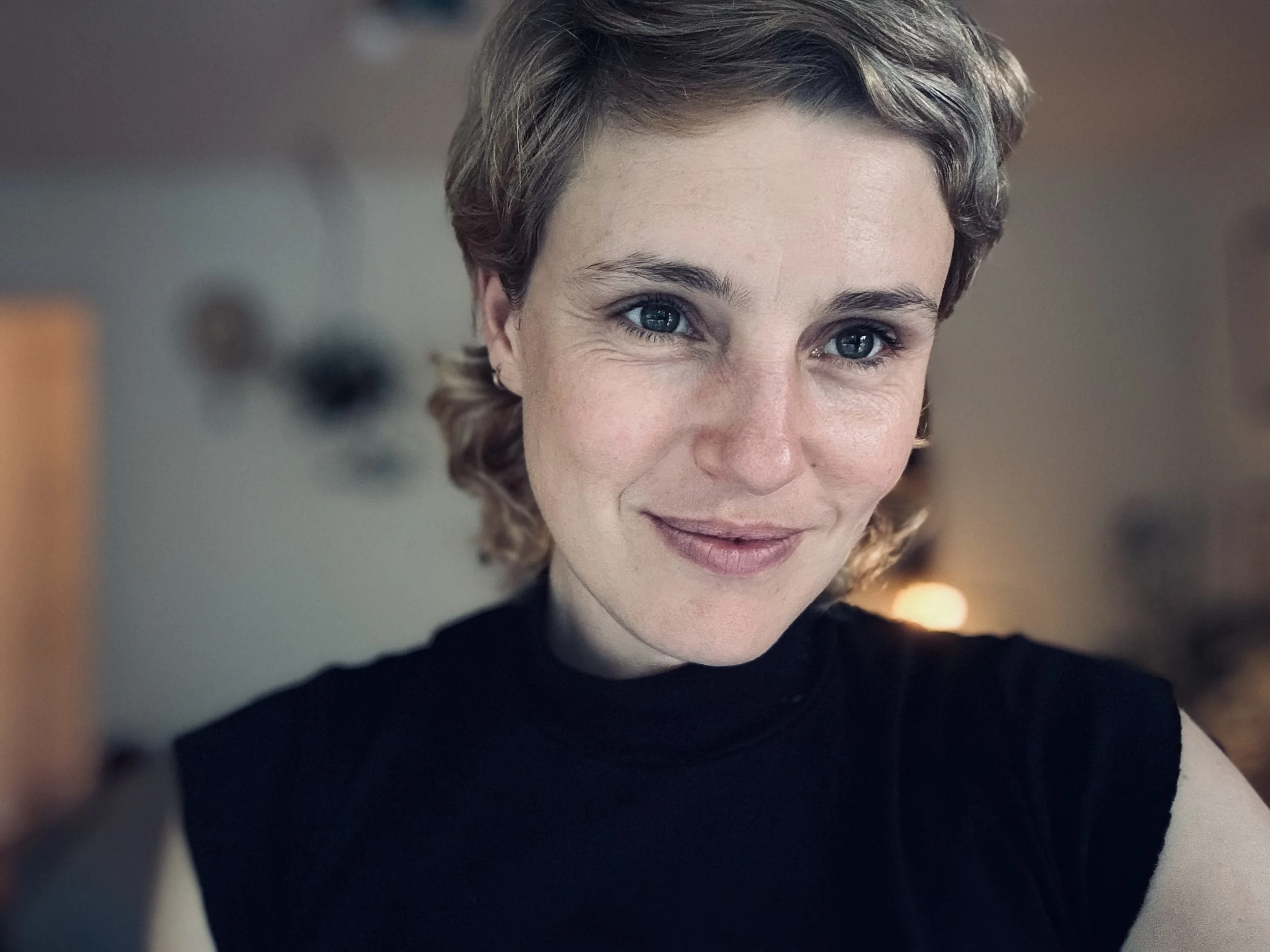 A woman with short, curly blond hair and blue eyes, wearing a black sleeveless top, smiling softly indoors.