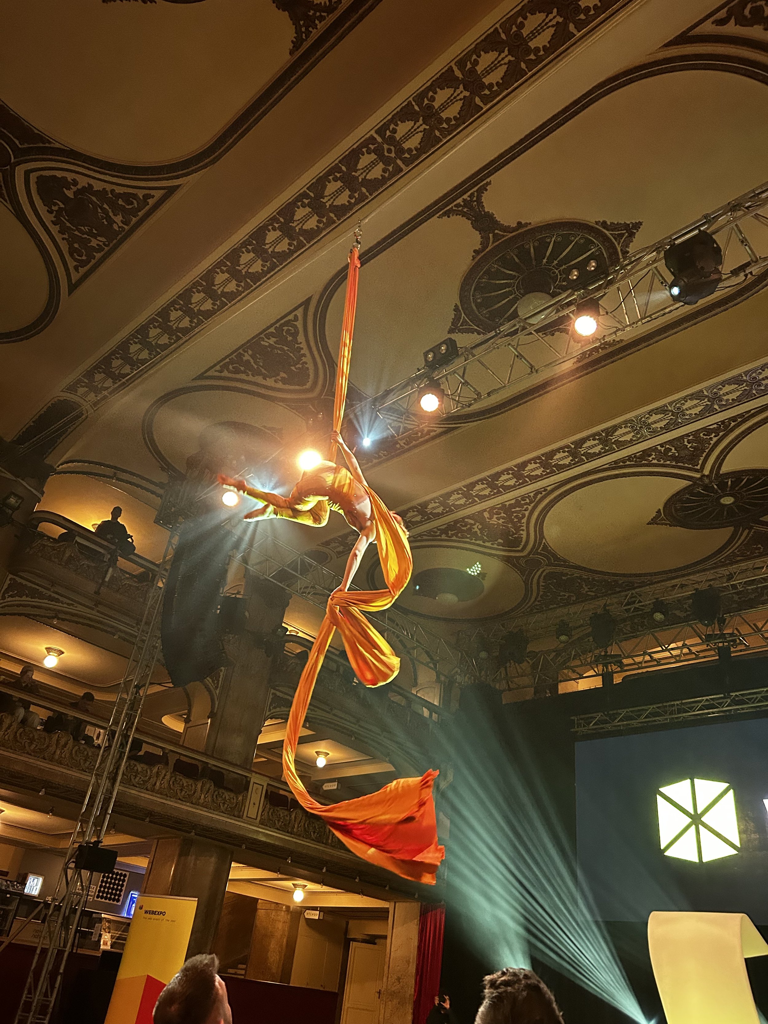 Ivy Kolcun an aerial silk performer suspended in mid-air during a stage show, with elaborate ceiling designs and stage lighting in the background performing at WebExpo 2025