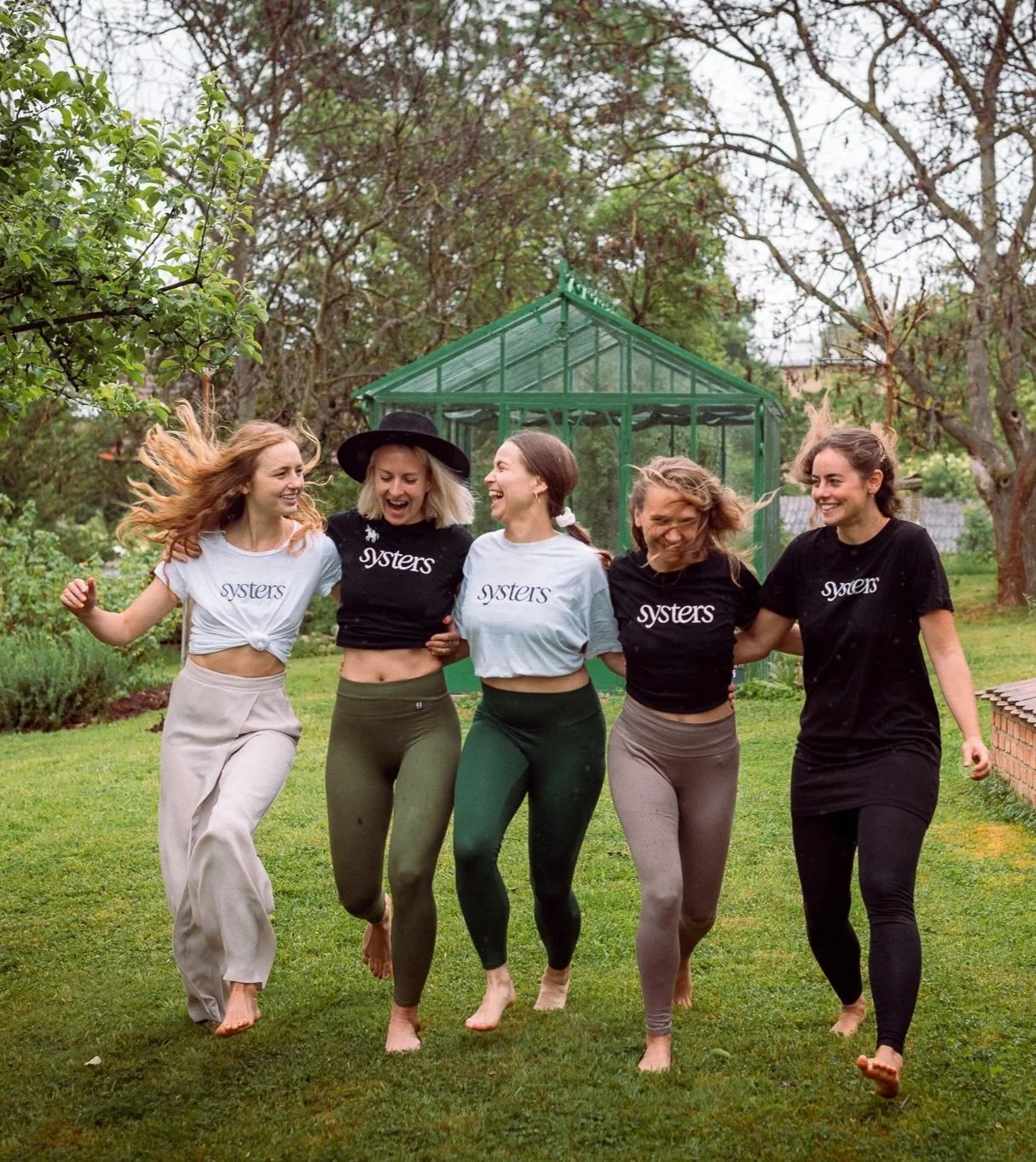 Five young women walking in a garden, smiling, with a greenhouse behind them.