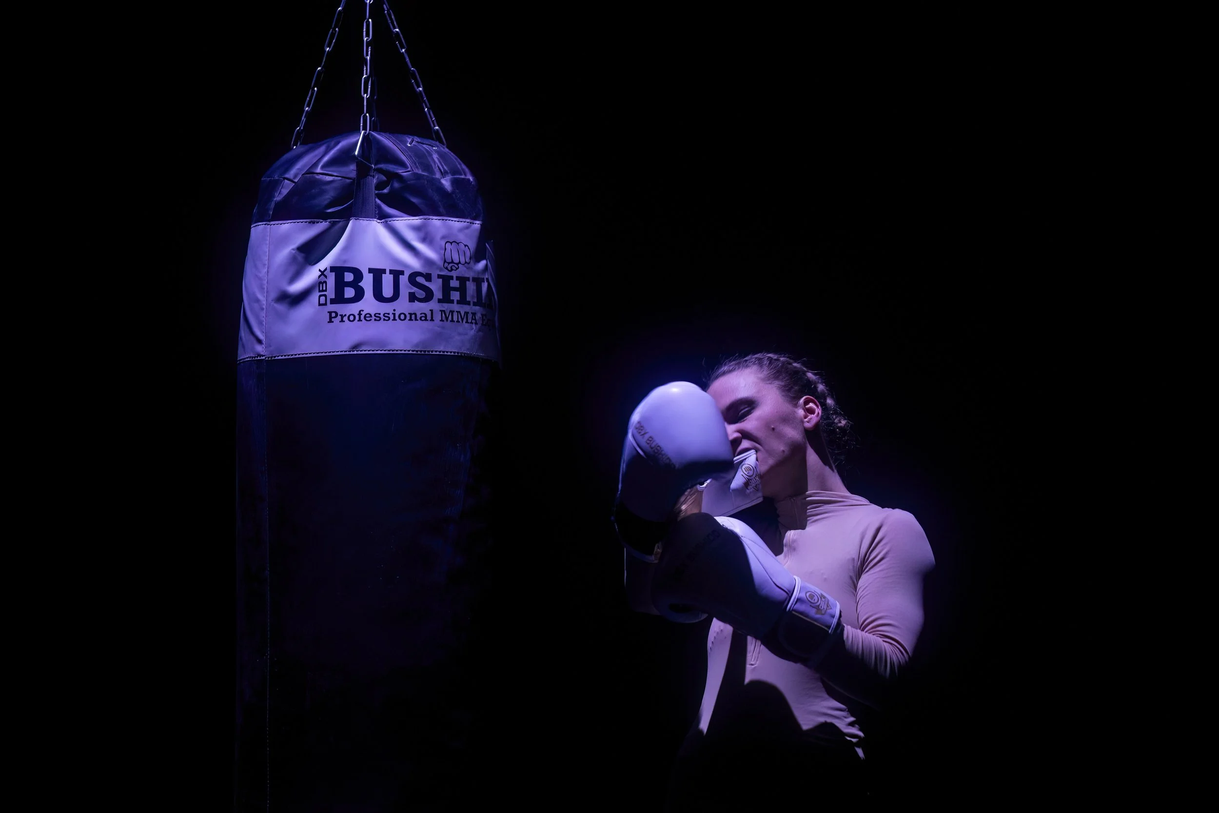 A female boxer in a beige long-sleeve top punches a hanging black and purple boxing bag labeled 'BUSHI Professional MMA' inside a dimly lit gym.