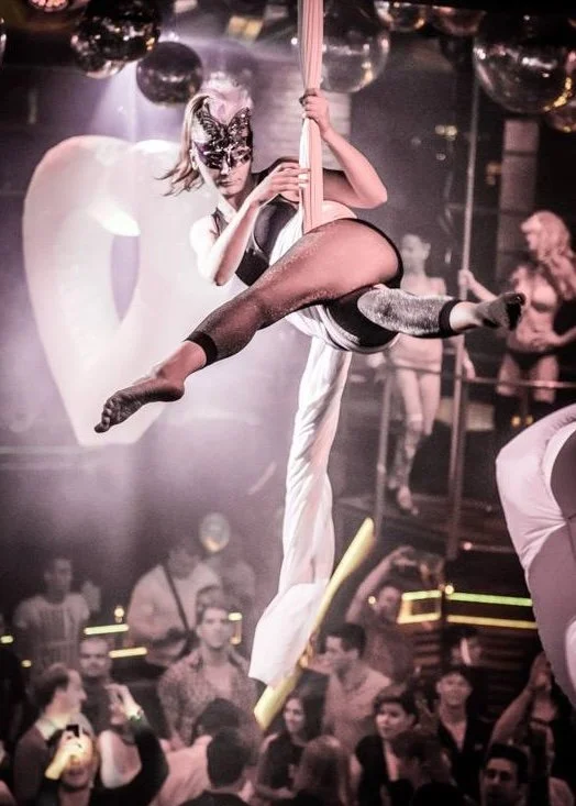 Ivy Kolcun an aerial silk performer dressed as a masked woman above a crowd in a nightclub, with other performers in the background.