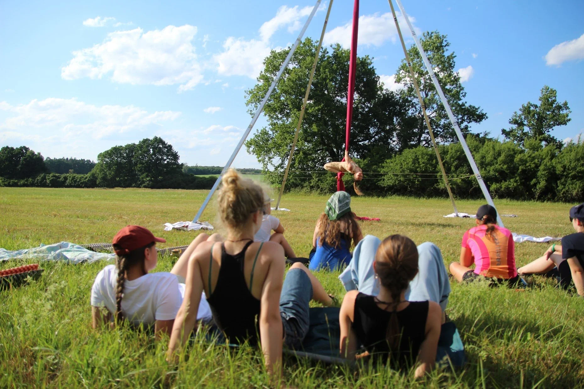 Group of children and teenagers sitting on grass in a field, watching a girl performing aerial silk acrobatics inside a large outdoor swing structure under a blue sky with clouds.