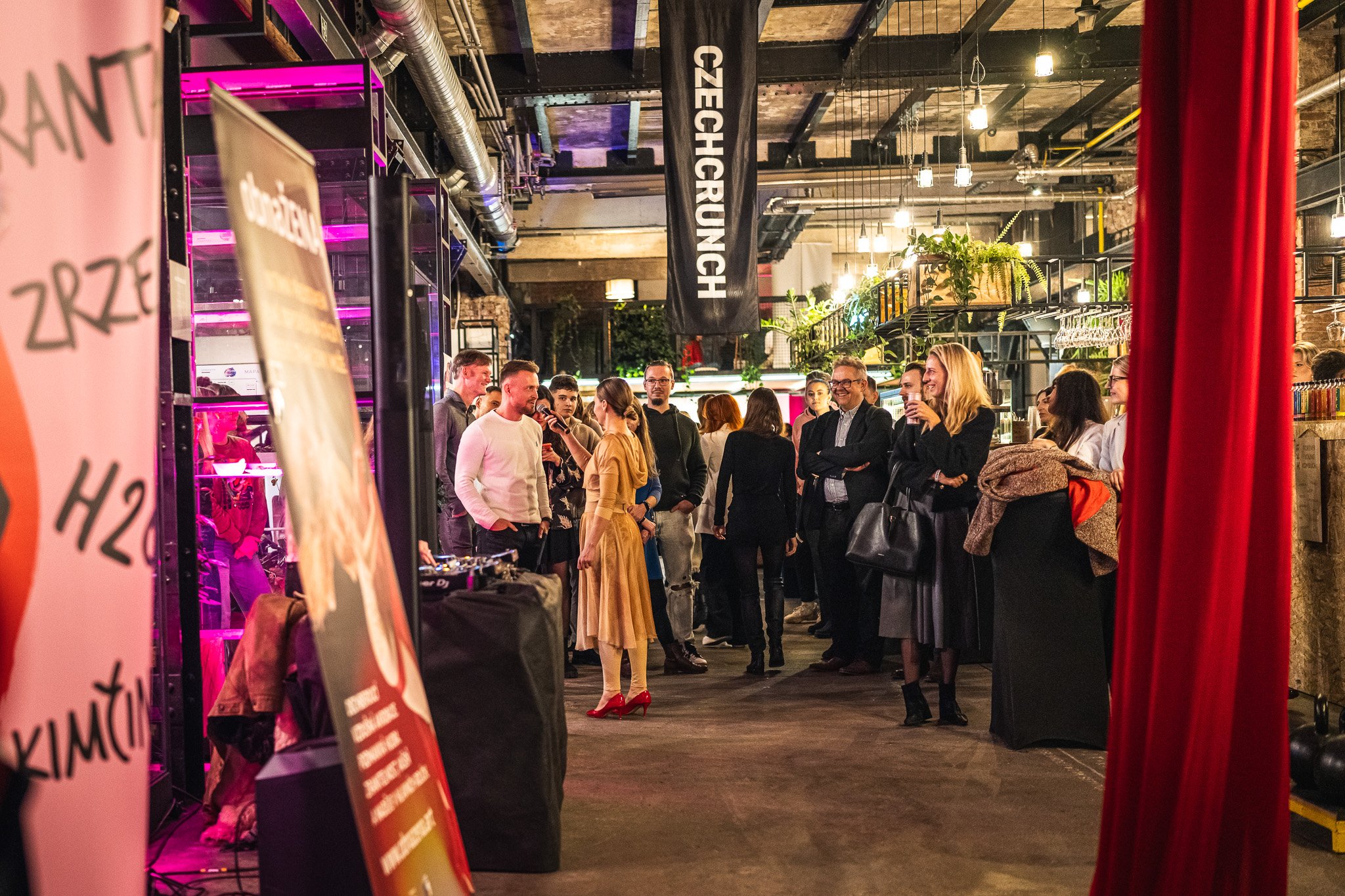 Group of people gathered in an industrial-style venue with exposed brick and ductwork, some talking and holding drinks, with a DJ setup visible on the left.