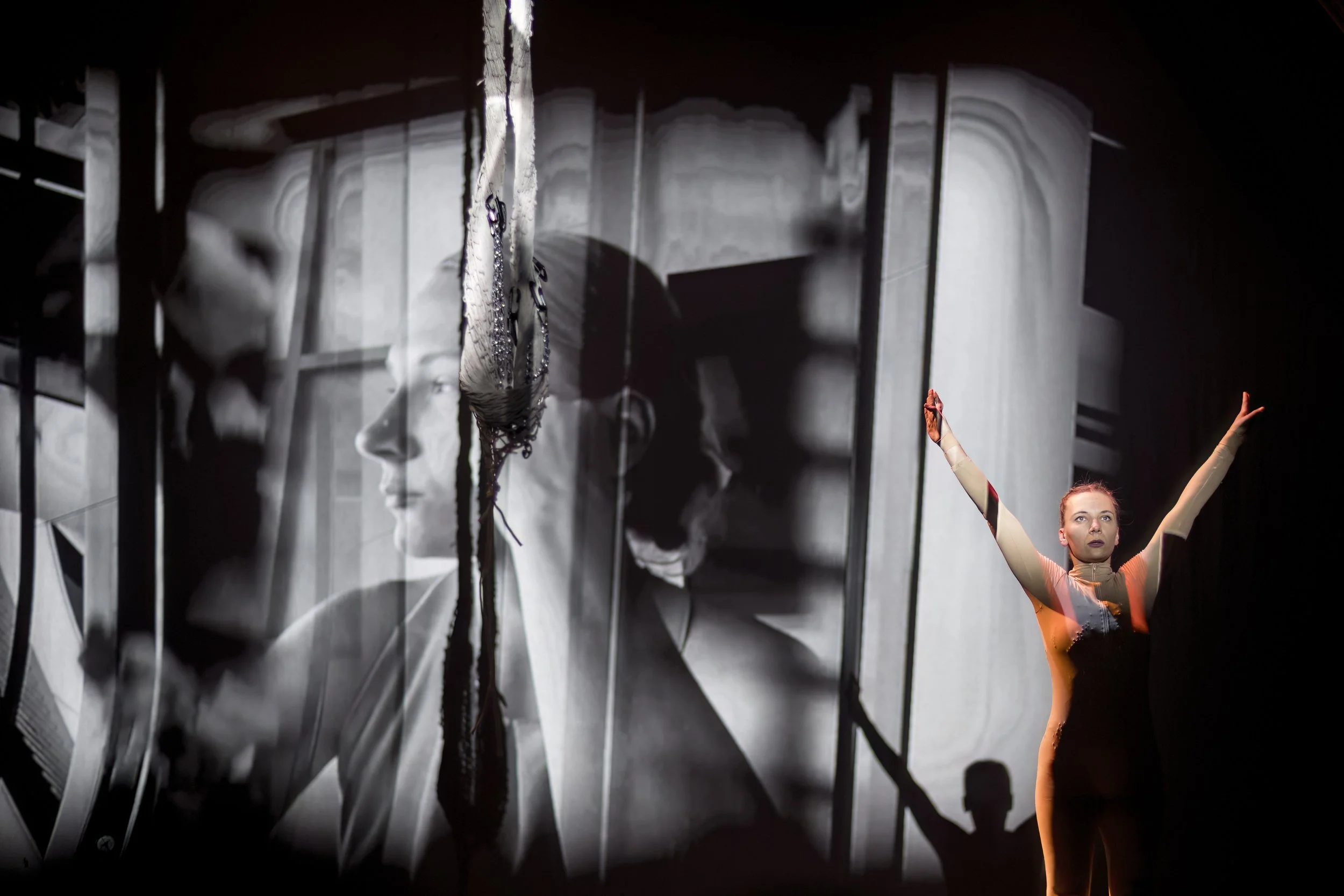 A performer with arms raised stands in front of a large black-and-white projection of a woman's face, with layered images and shadows creating an artistic visual effect.