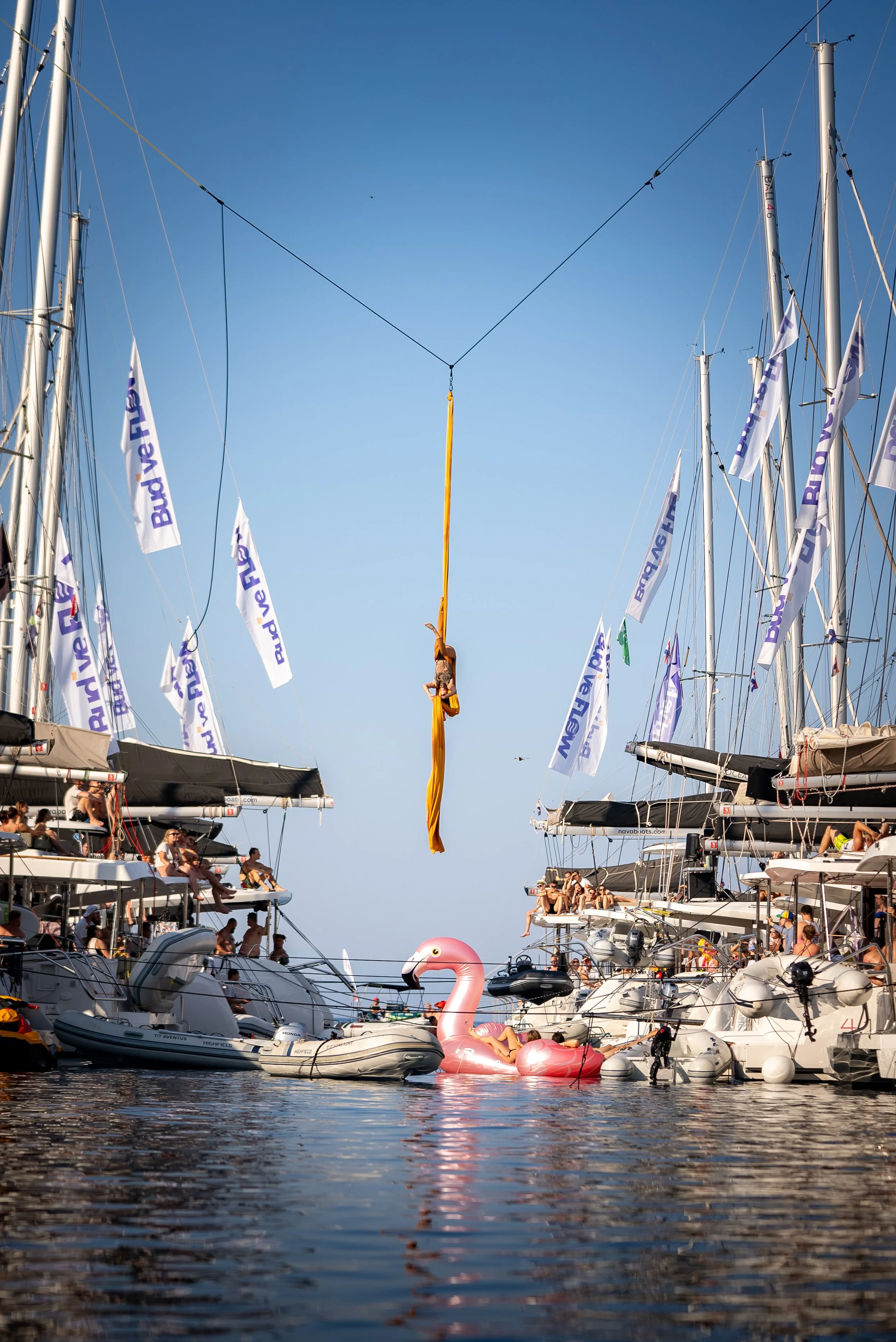 Ivy Kolcun an aerial silk performer hanging upside down on yellow fabric between two sailboats at a marina with many boats and people, a blue sky, and a floating pink flamingo as a part of festival FLOW 2025
