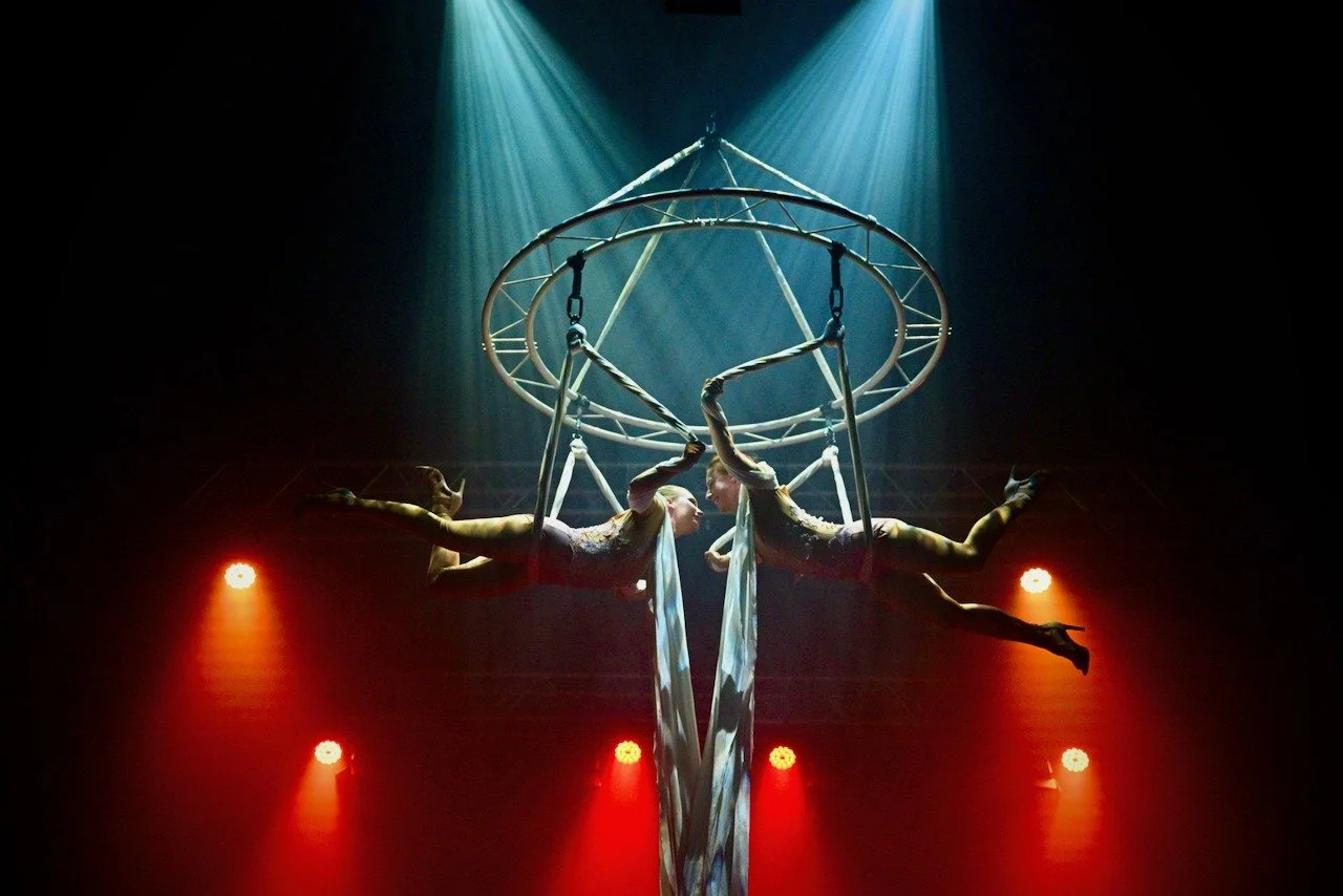 Two aerial performers in glittery costumes perform on hanging apparatus under spotlights, creating a symmetrical pose with their bodies facing each other during a show.