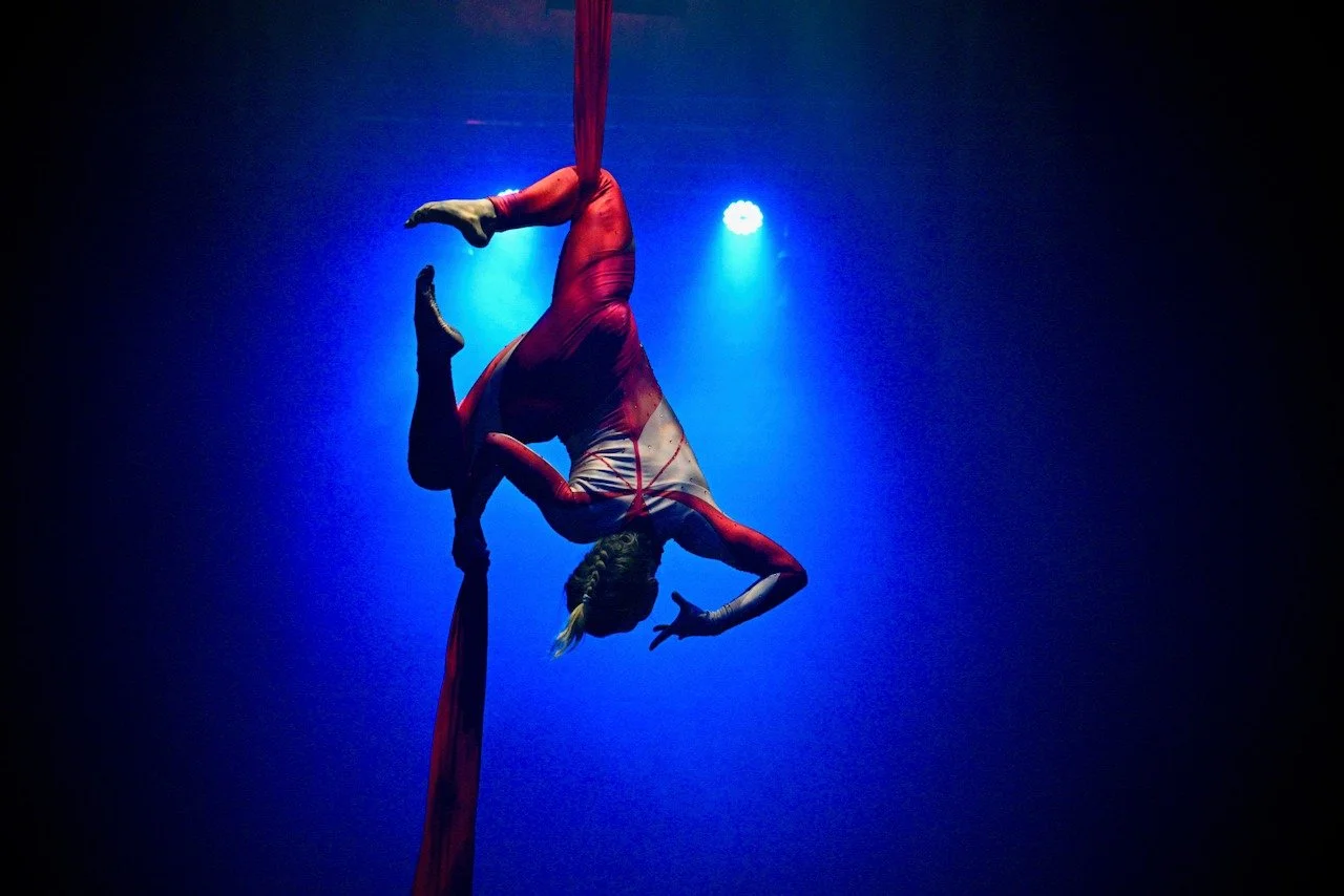 Circus performer dressed in red and white costume hanging upside down on aerial silk fabric against dark background with blue light highlighting the act.