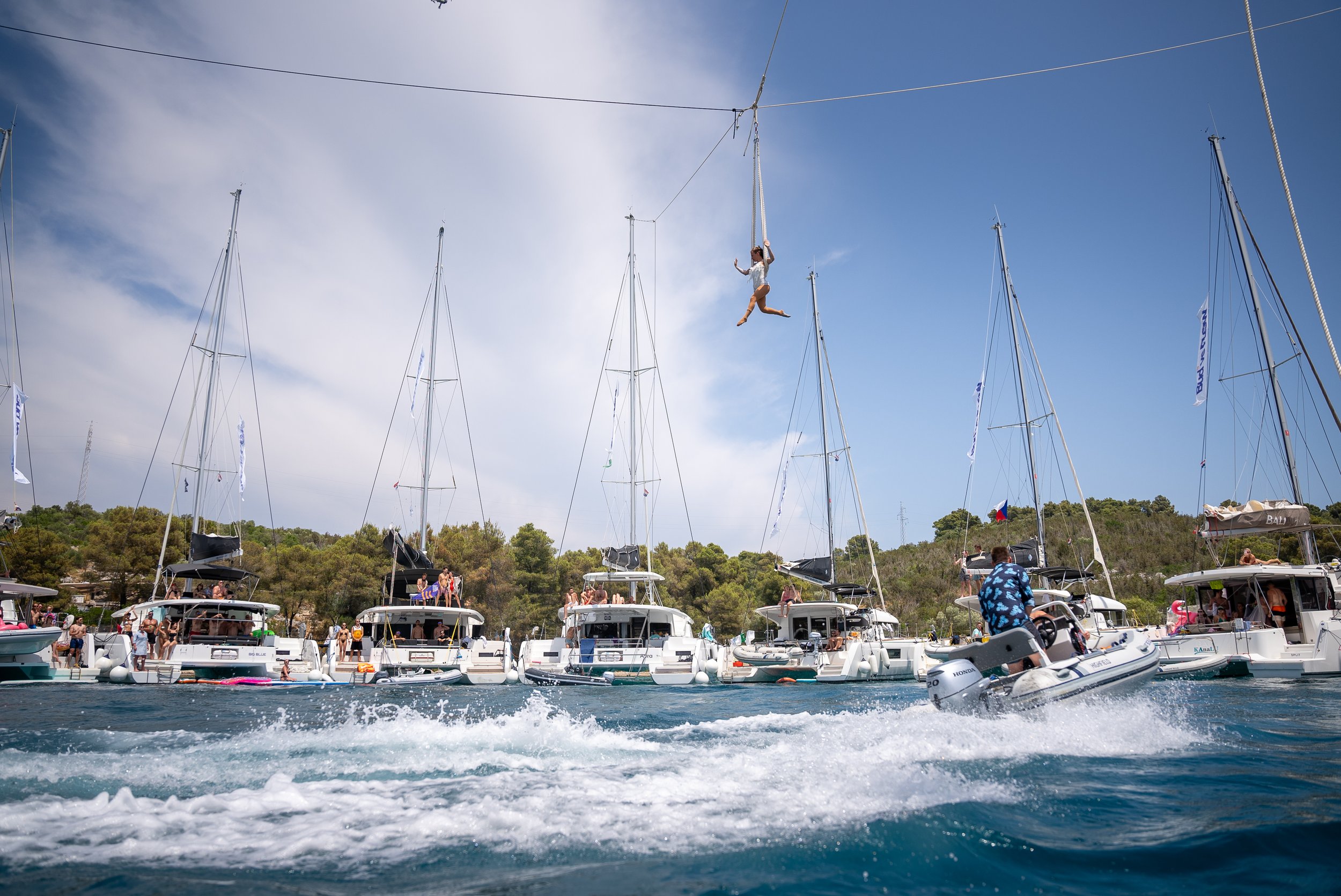 Ivy Kolcun an aerial silk performer performs an aerial silk stunt above a marina with multiple boats and yachts, with people on deck and a person on a small boat below, all under a partly cloudy sky festival FLOW 2025