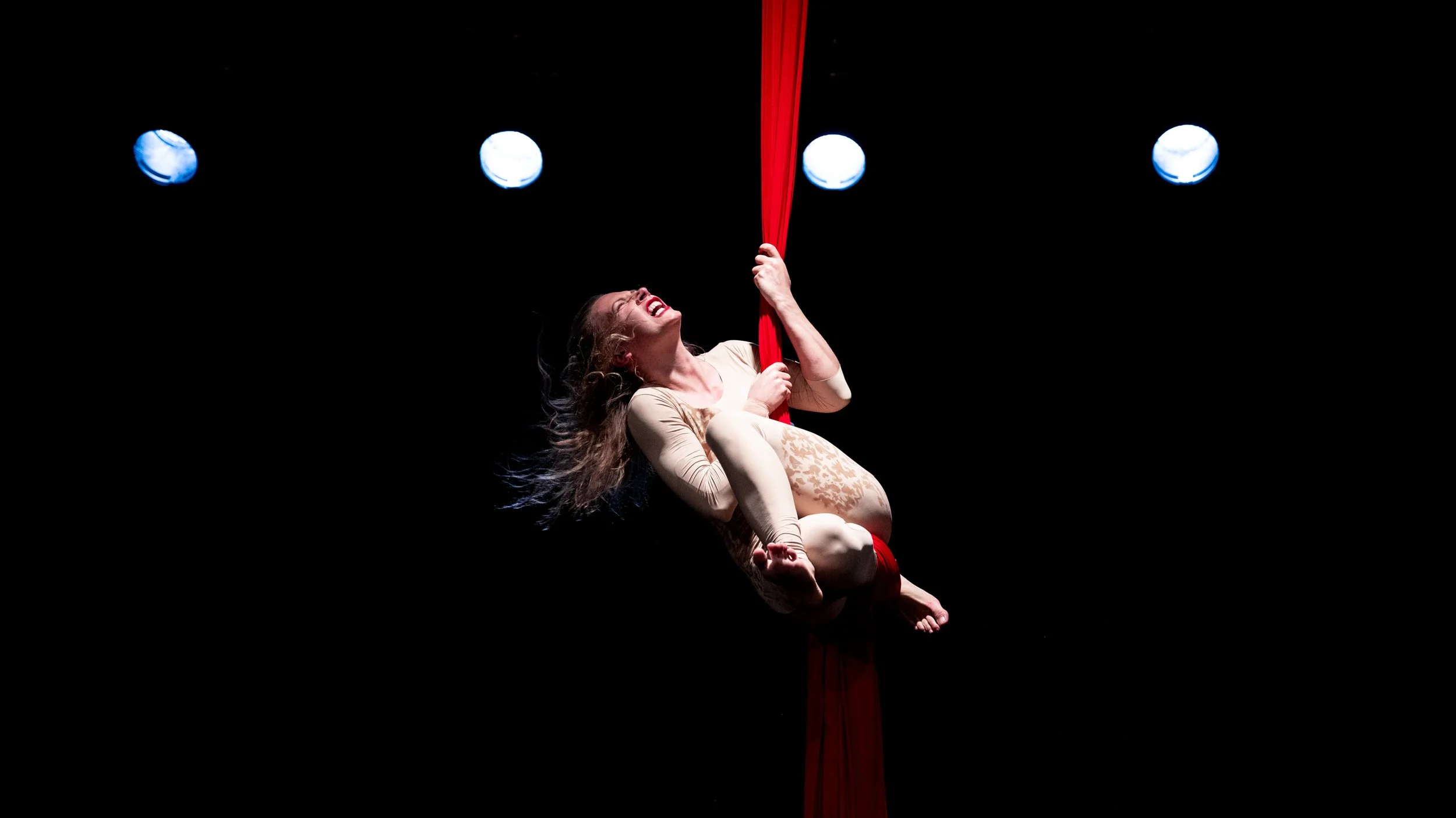 A woman performing an aerial silk routine on a dark stage with three spotlights overhead, she is smiling and holding onto a red silk fabric.