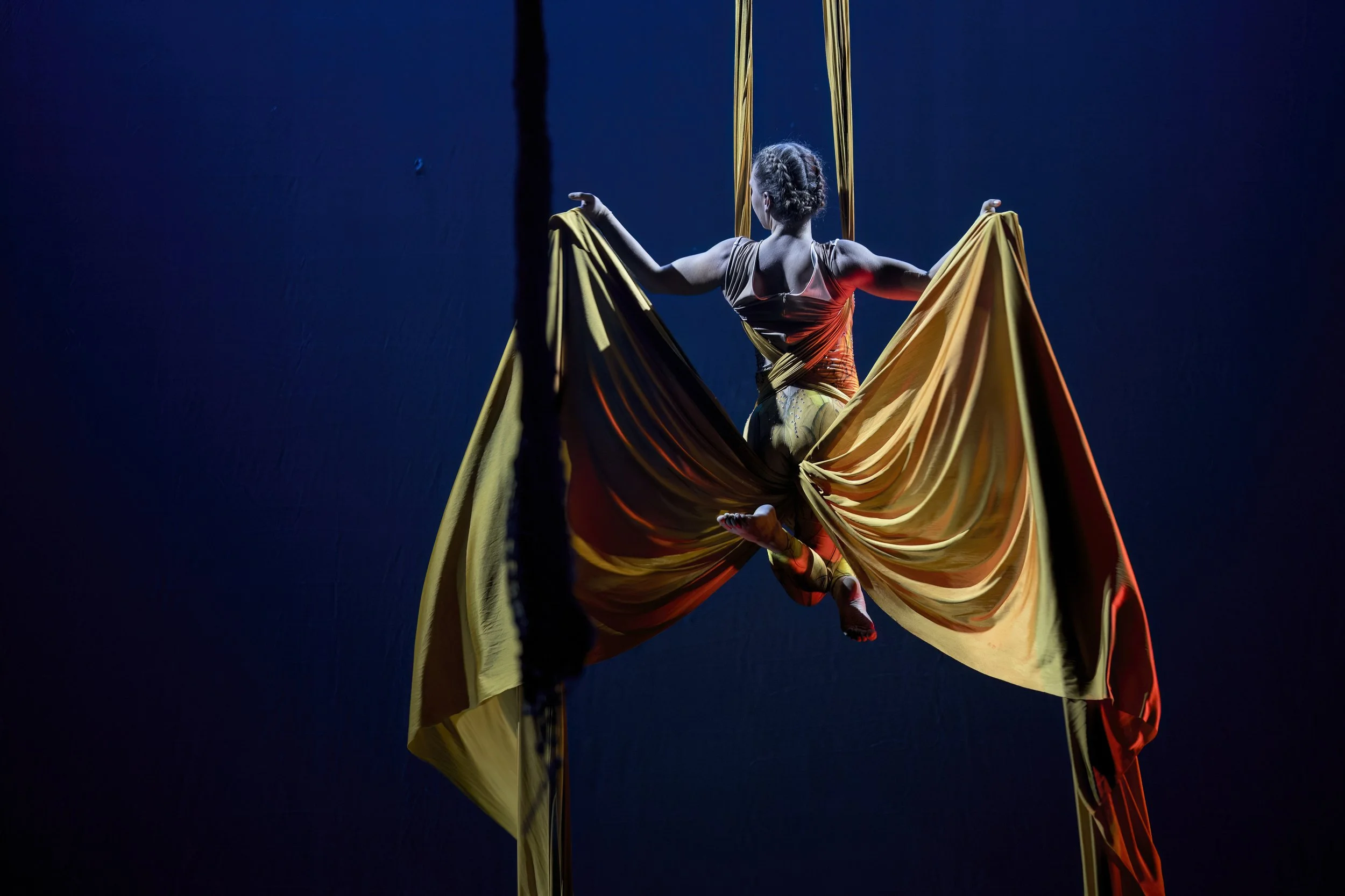 Aerial silk performer suspended in the air, holding yellow and orange silk fabric against a dark blue background.