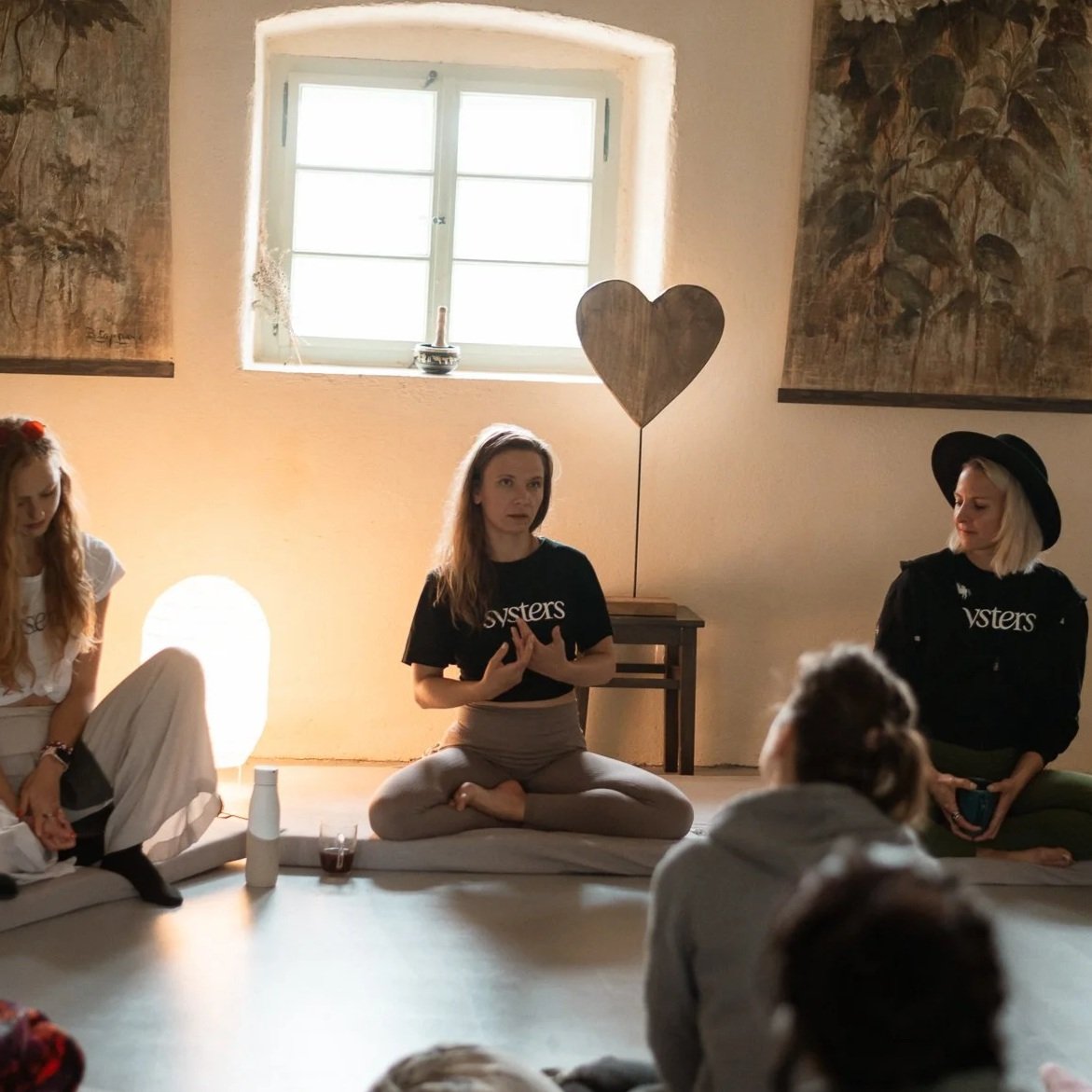 A group of women sitting on the floor inside a room with a window, engaged in a discussion or meditation session. One woman in the center is speaking and making hand gestures, with others listening. There is a large wooden heart sculpture on a table 