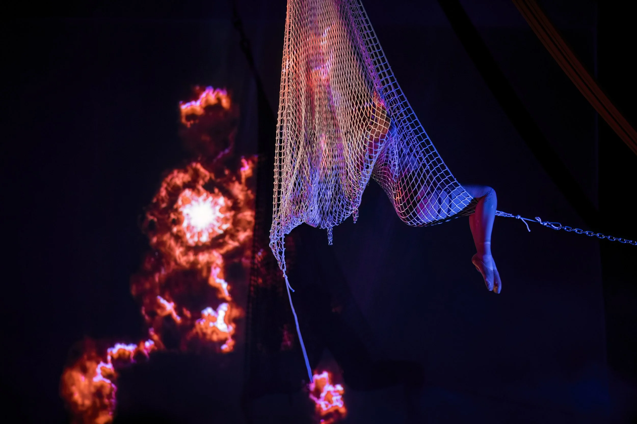An art installation featuring a suspended mannequin or figure wrapped in a white netted material, with colorful lighting, against a dark background with fiery abstract visuals.