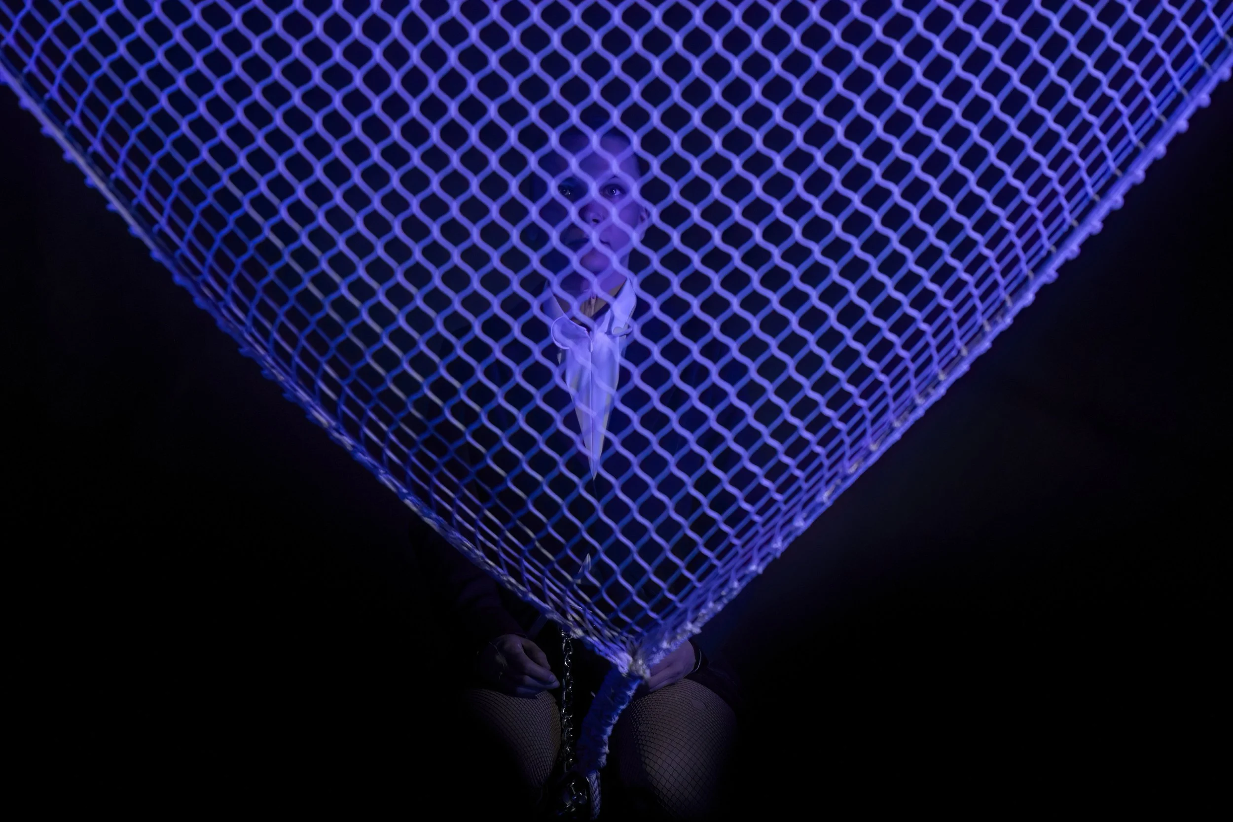 A person sitting under a large, purple-lit wireframe pyramid, with only the face and hands visible against a dark background.