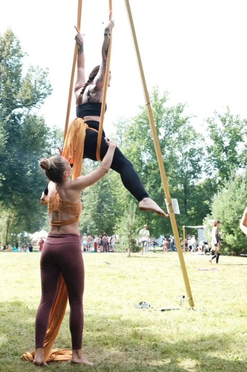 Aerial silk performers practicing outdoors in a park, with one performer hanging upside down from yellow aerial silks and the other supporting her from below.
