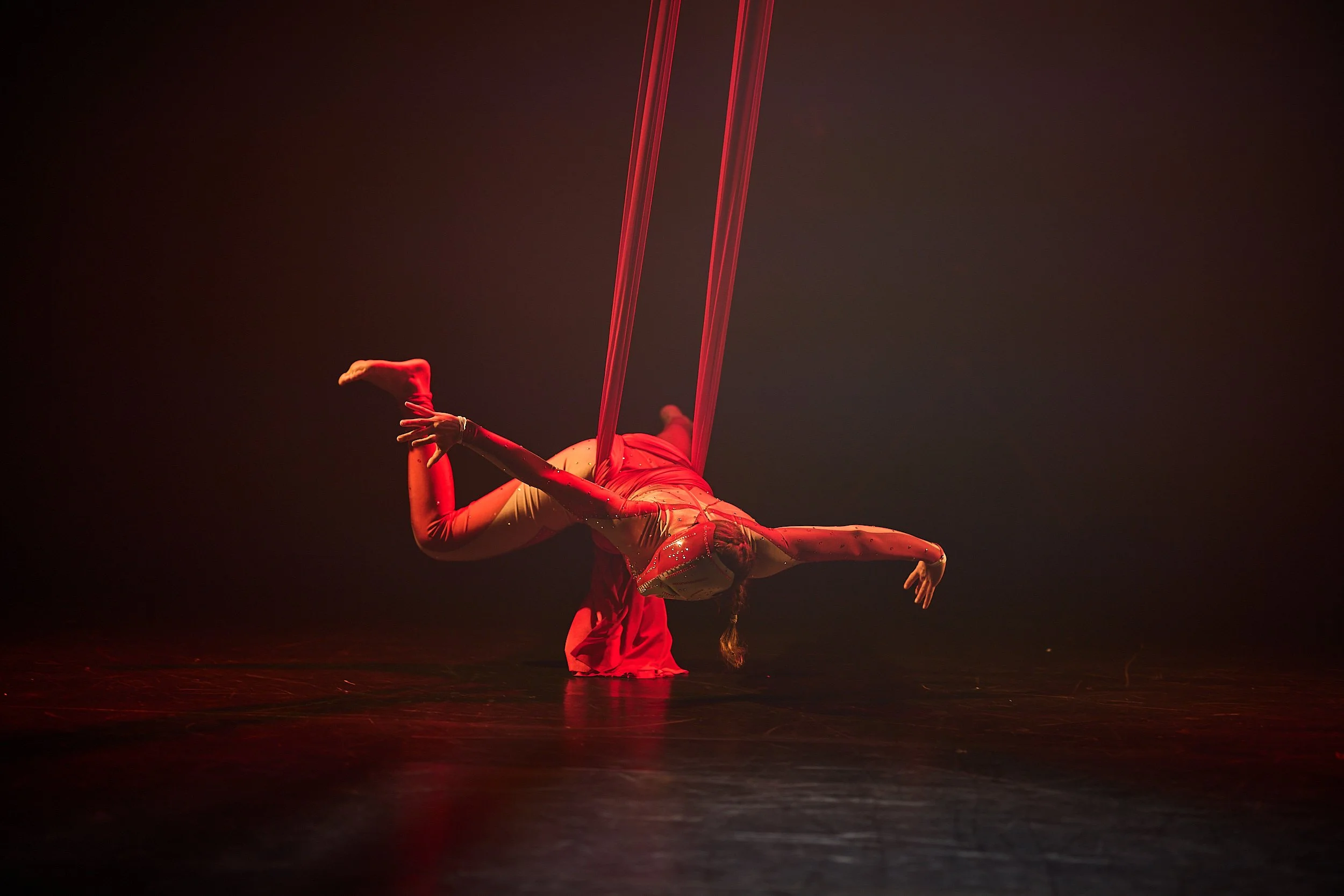 A performer in a red costume is suspended upside down in a dance or acrobatics routine, using red silk fabric for aerial silk artistry on a dark stage.