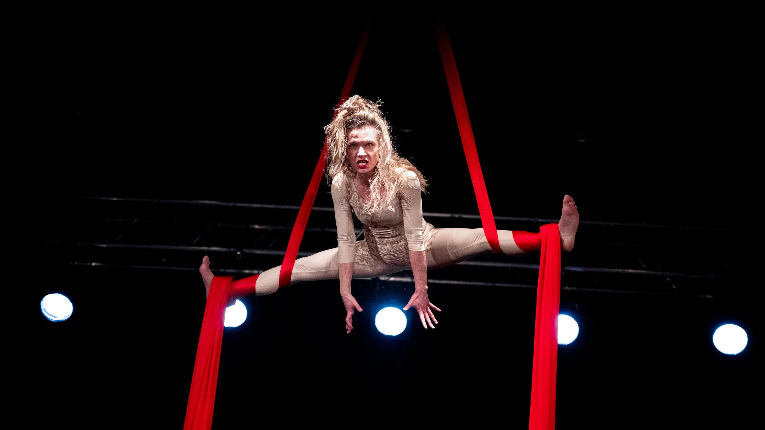 A female aerialist performing an upside-down split on red aerial silk fabric, with an intense facial expression, against a dark background and stage lights.