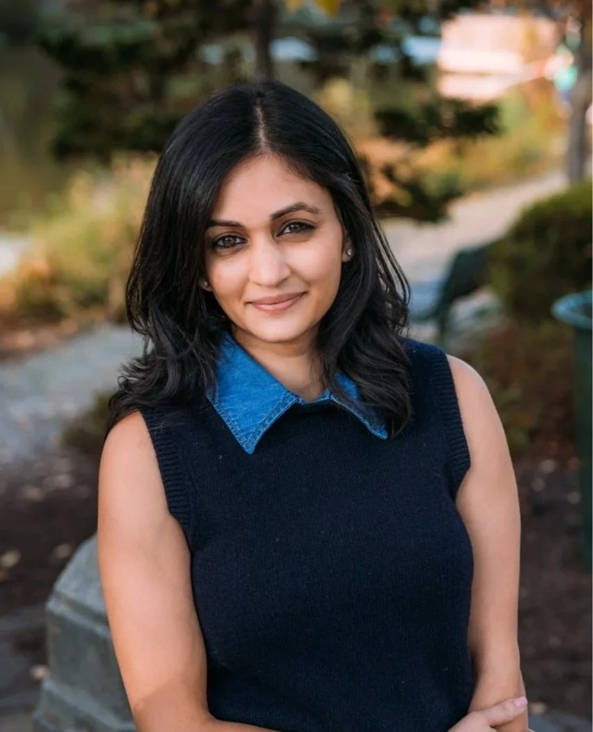 A woman with medium skin tone, black hair, and a gentle smile, standing outdoors with trees and a clothing rack in the background.