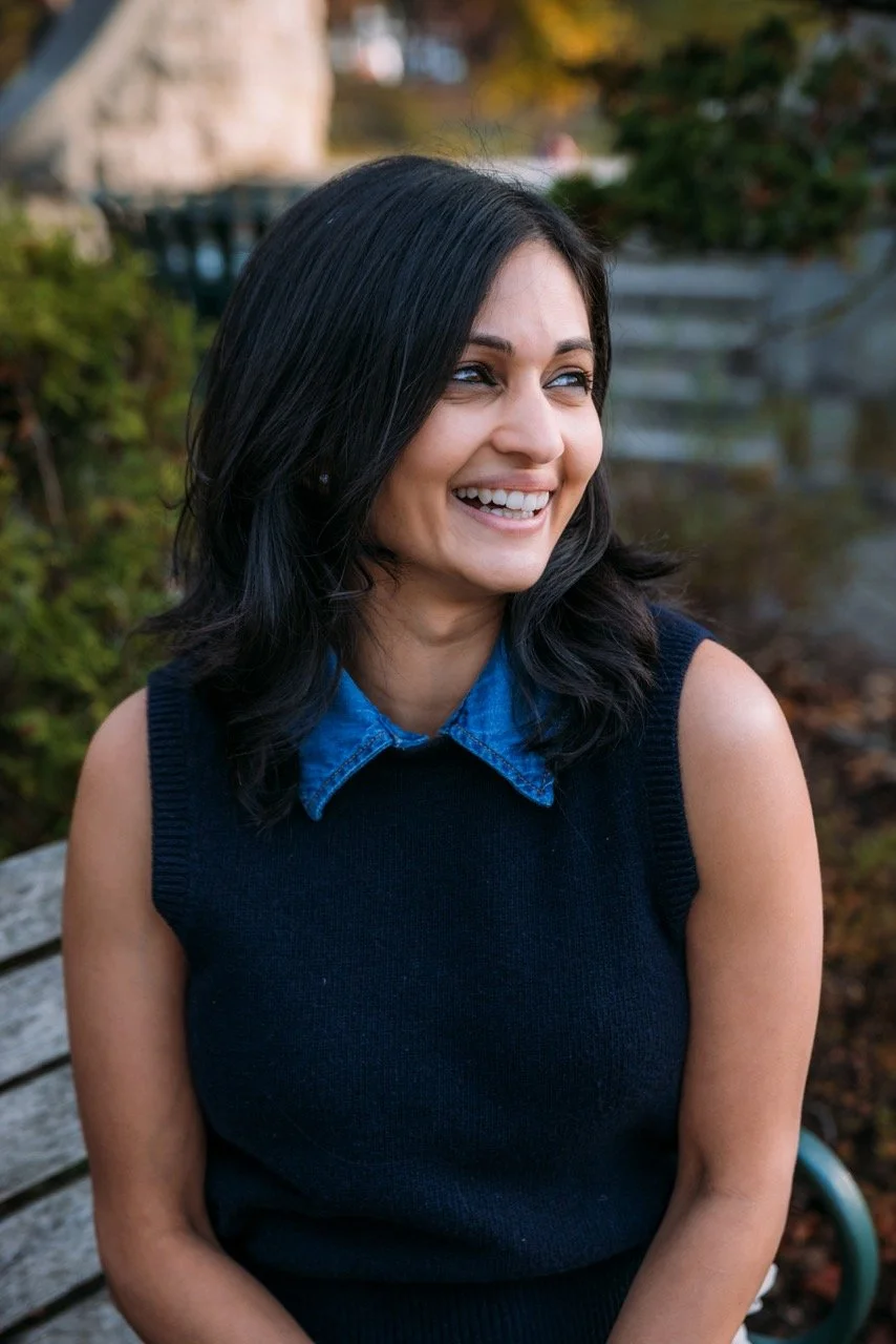 A woman with black hair and a denim shirt layered under a sleeveless black sweater, smiling outdoors near water and greenery.