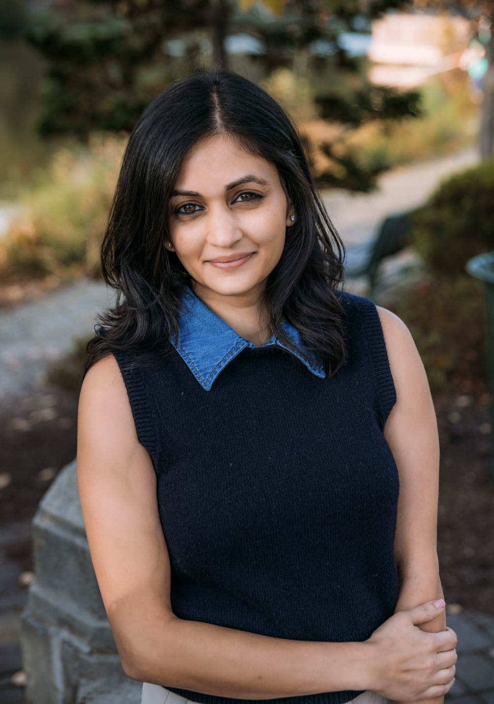 A woman with dark hair and a slight smile, wearing a denim shirt under a black sleeveless sweater, standing outdoors in a park with trees and benches in the background.