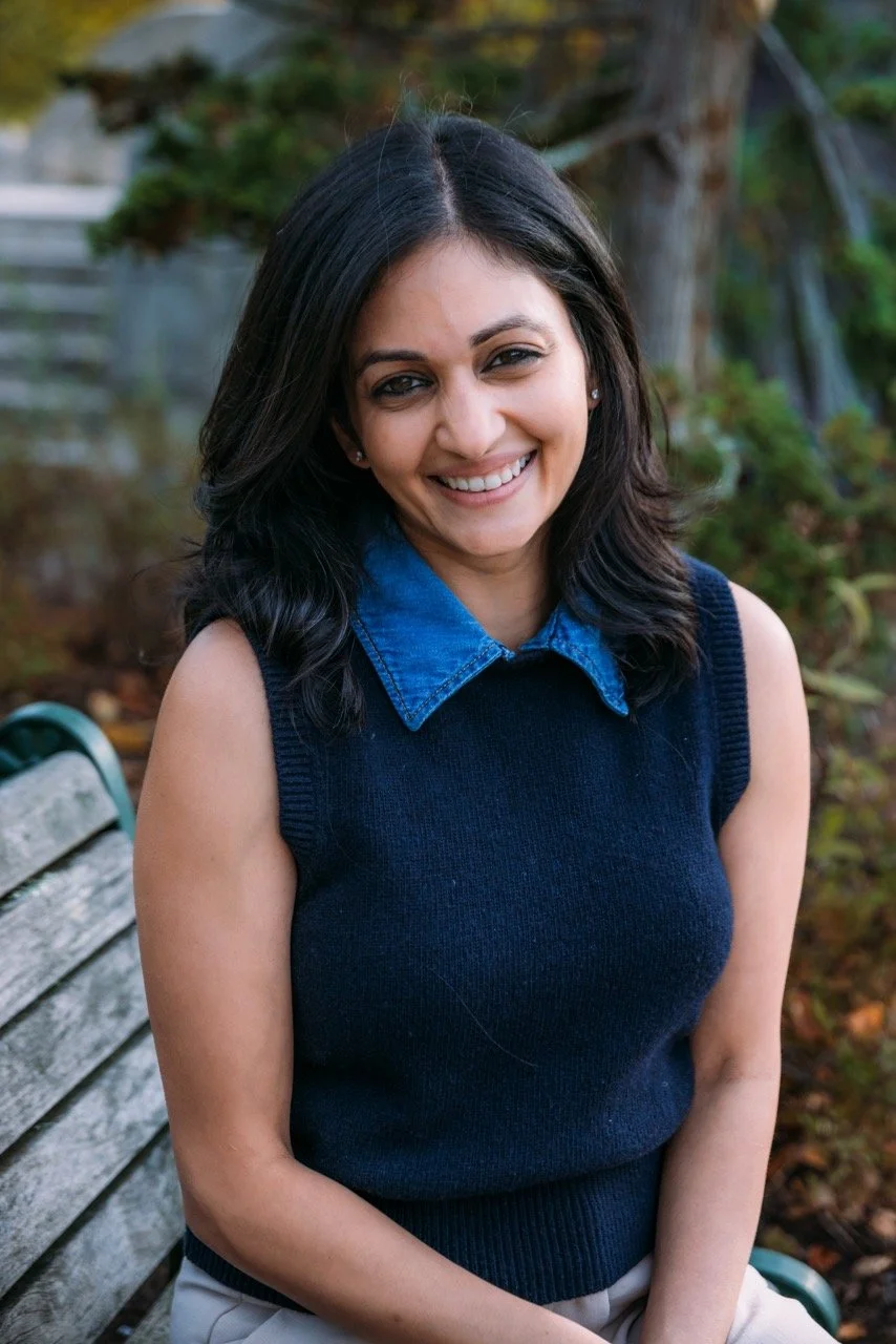 A woman with shoulder-length black hair, wearing a sleeveless navy blue sweater over a denim shirt, smiling while sitting on a park bench outdoors.
