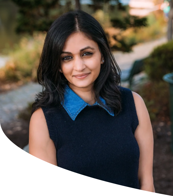 A woman with shoulder-length black hair, wearing a sleeveless black sweater over a blue collared shirt, standing outdoors with greenery and trees in the background.