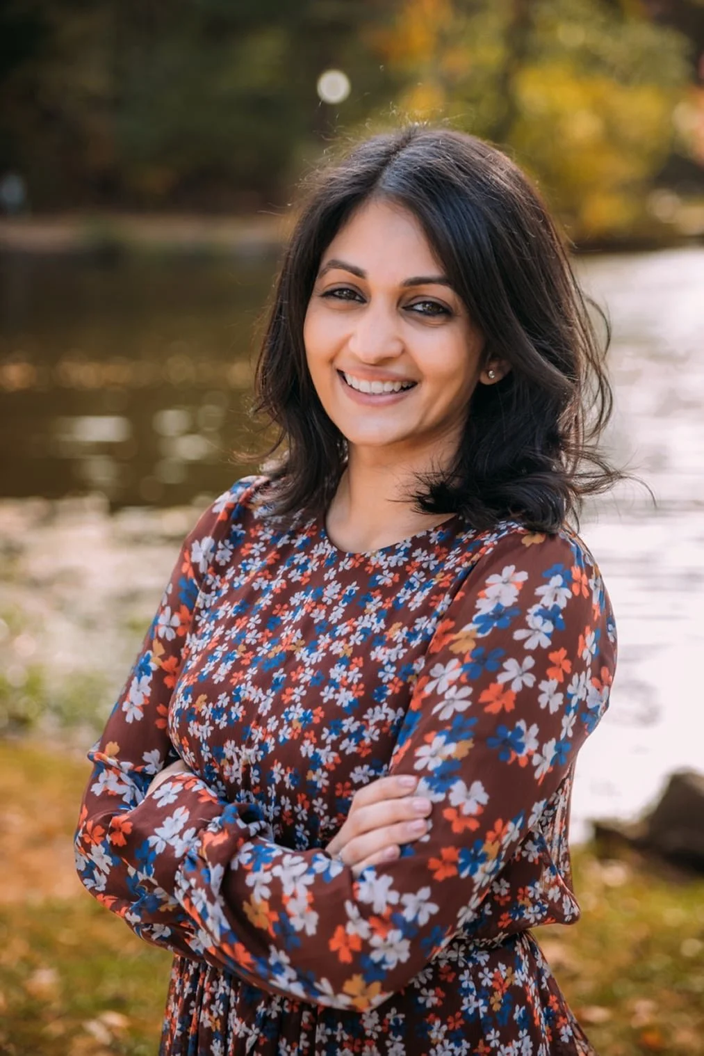 A woman with dark hair smiling outdoors near a river, wearing a brown dress with a colorful floral pattern, autumn trees in the background.