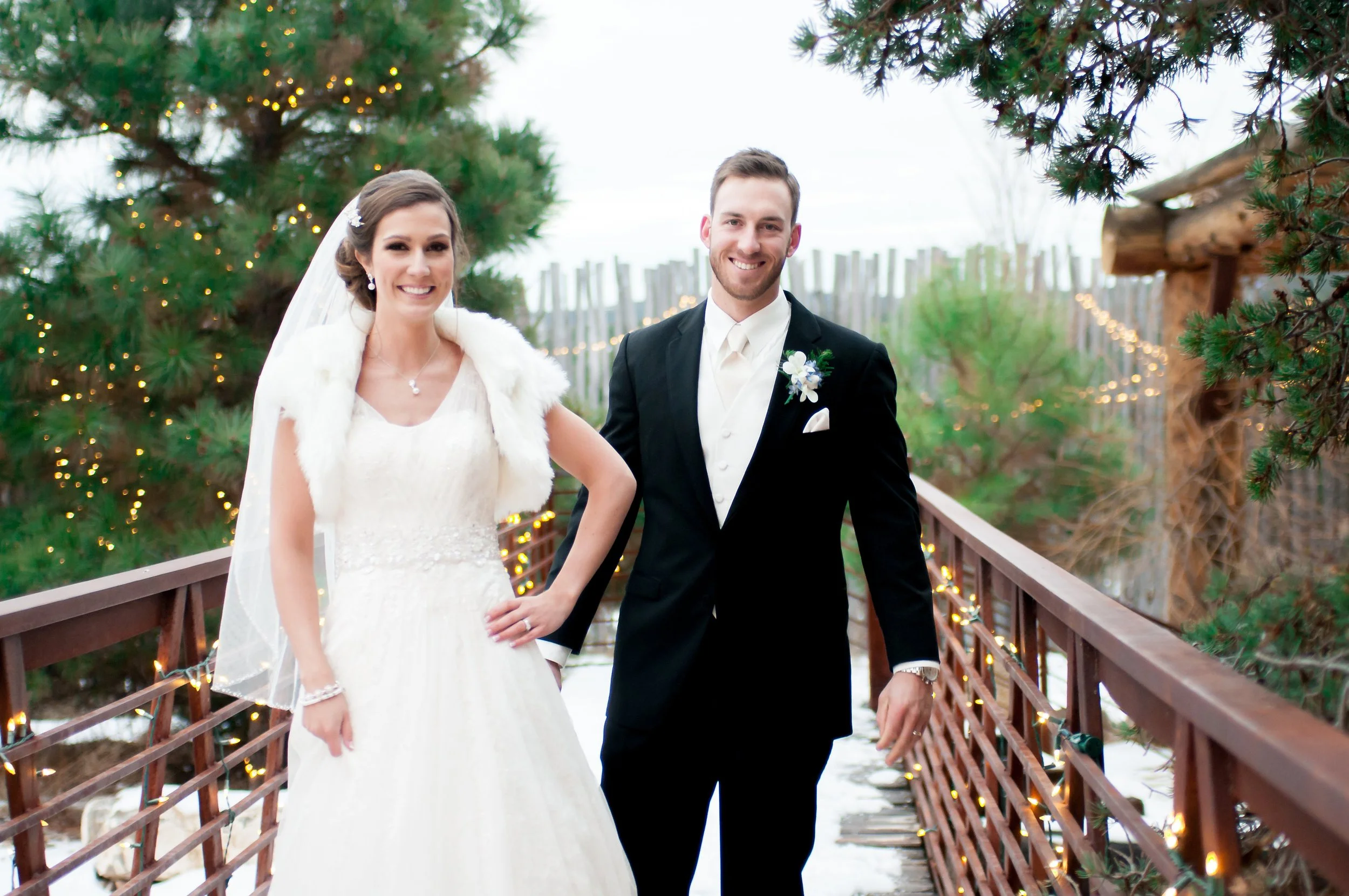 A bride and groom standing on a wooden bridge outdoors, smiling at the camera, with Christmas lights and evergreen trees in the background.