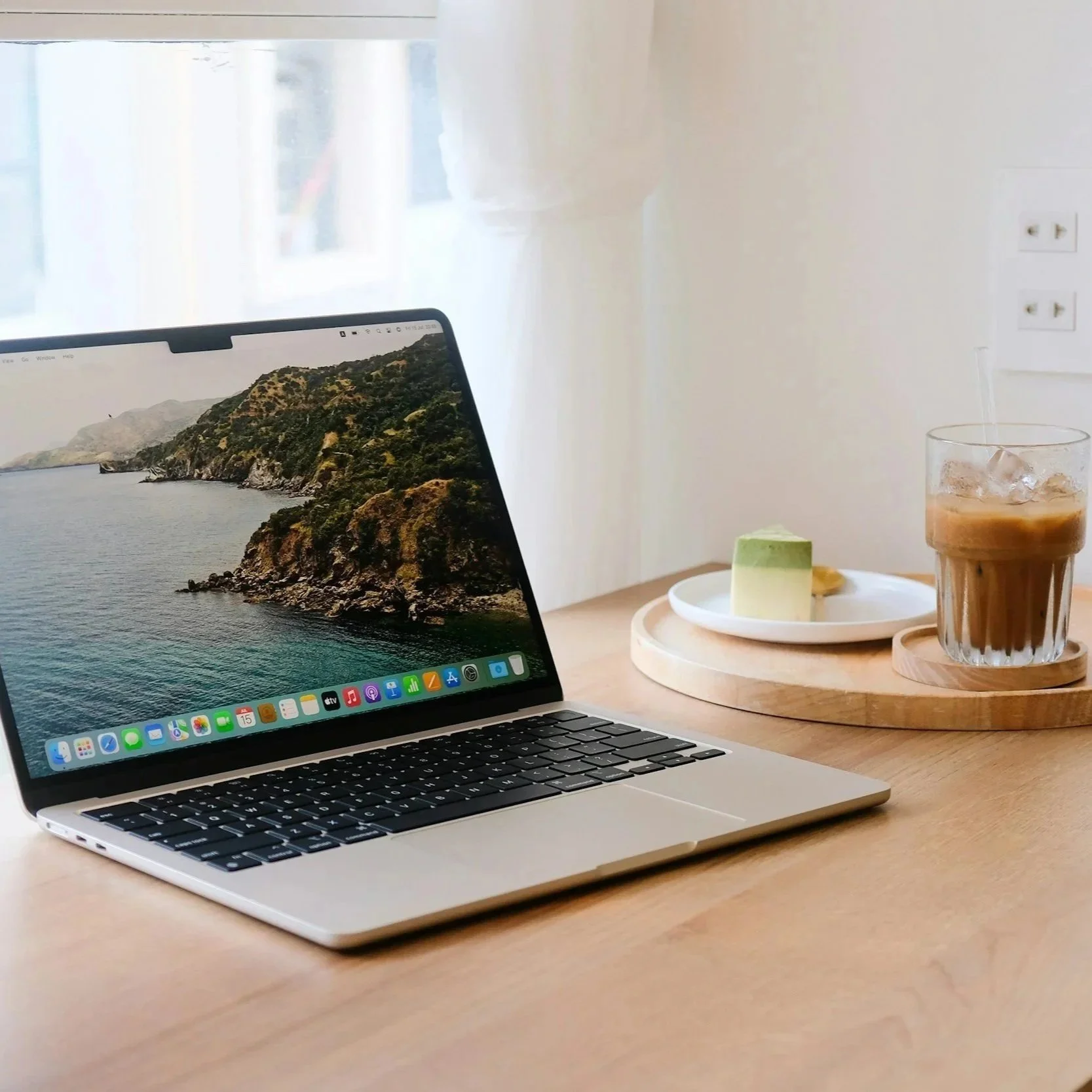 A laptop on a wooden desk with a coastal landscape wallpaper, next to a plate with a green dessert and a cookie, and a glass of iced coffee on a round wooden tray in a bright room.