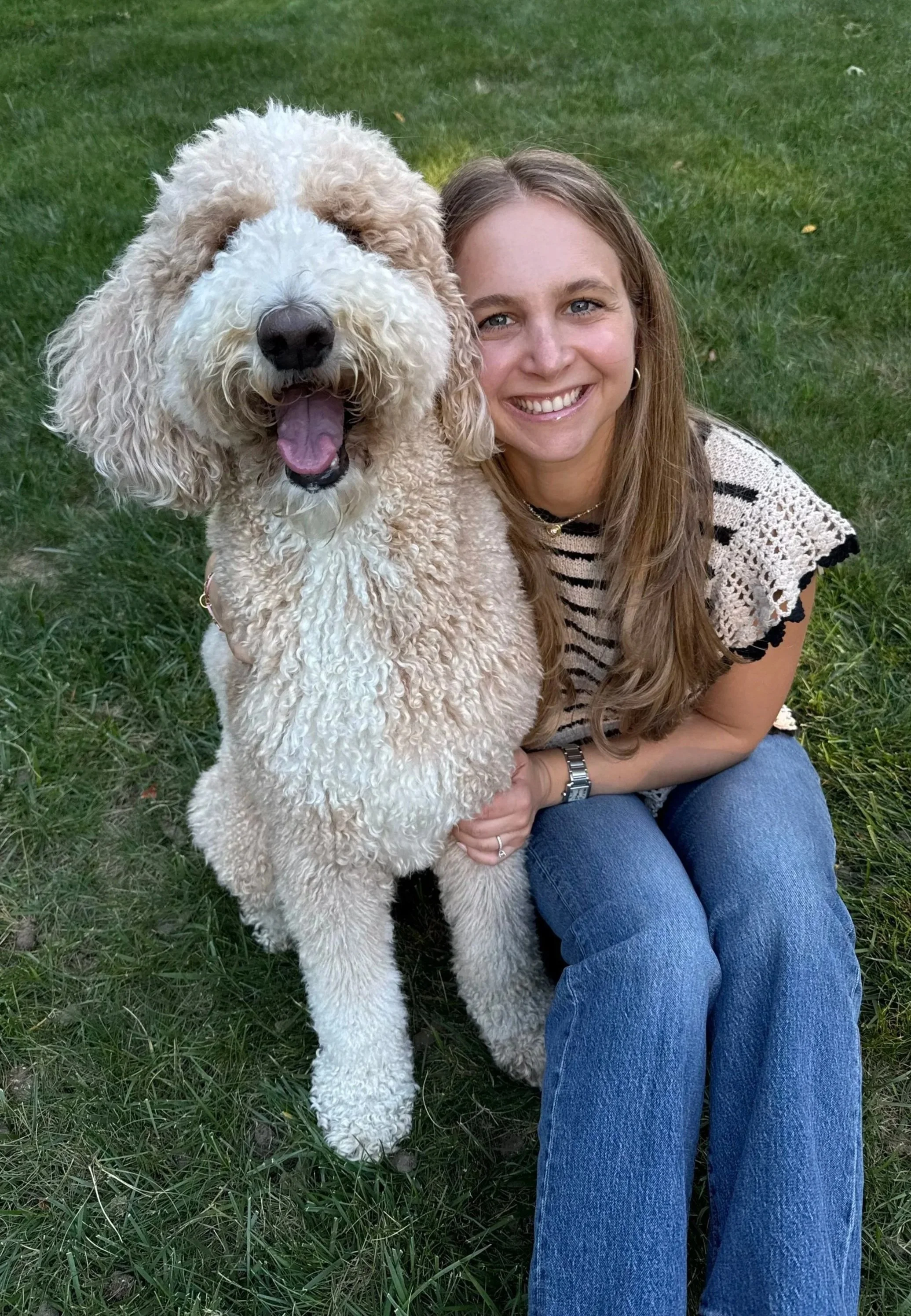 A woman with long, wavy hair, wearing a striped top and blue jeans, smiling while sitting on the grass next to a large, curly-haired dog with light-colored fur.