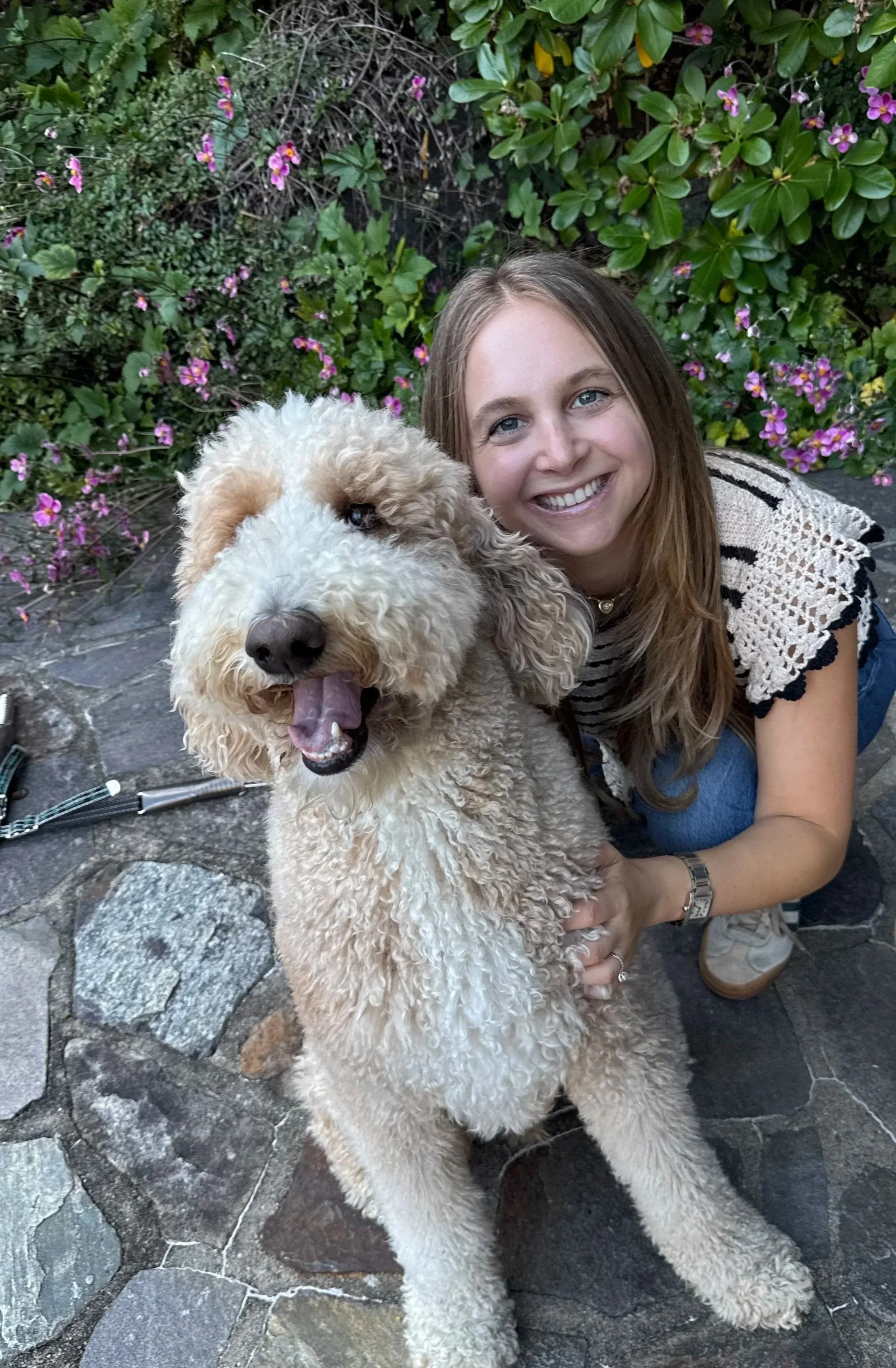 A young woman with long brown hair smiling and hugging a large, curly-coated beige dog with a happy expression in a garden with pink and purple flowers.