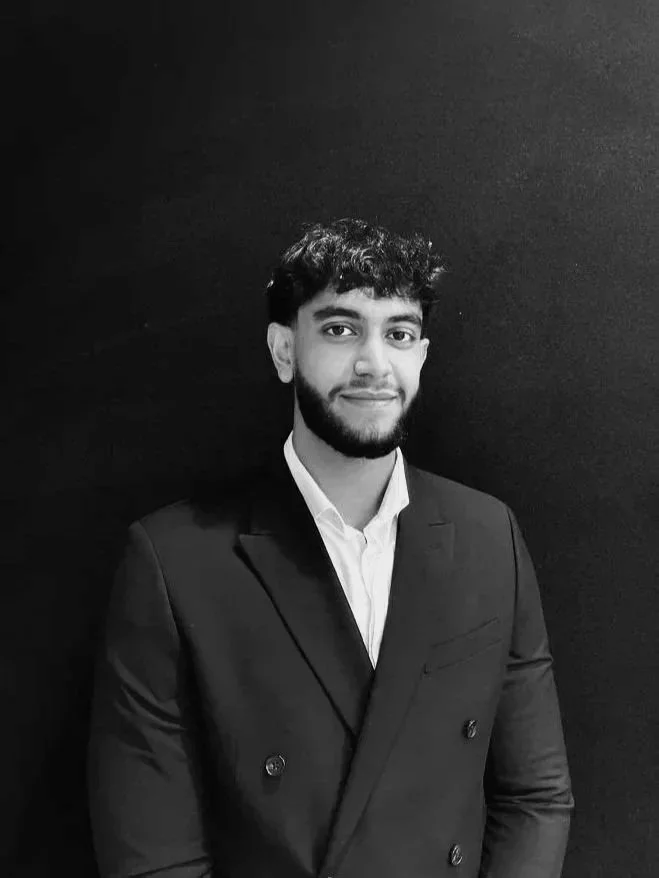 A young man with curly hair and a beard, wearing a suit and white shirt, standing against a dark background and smiling.