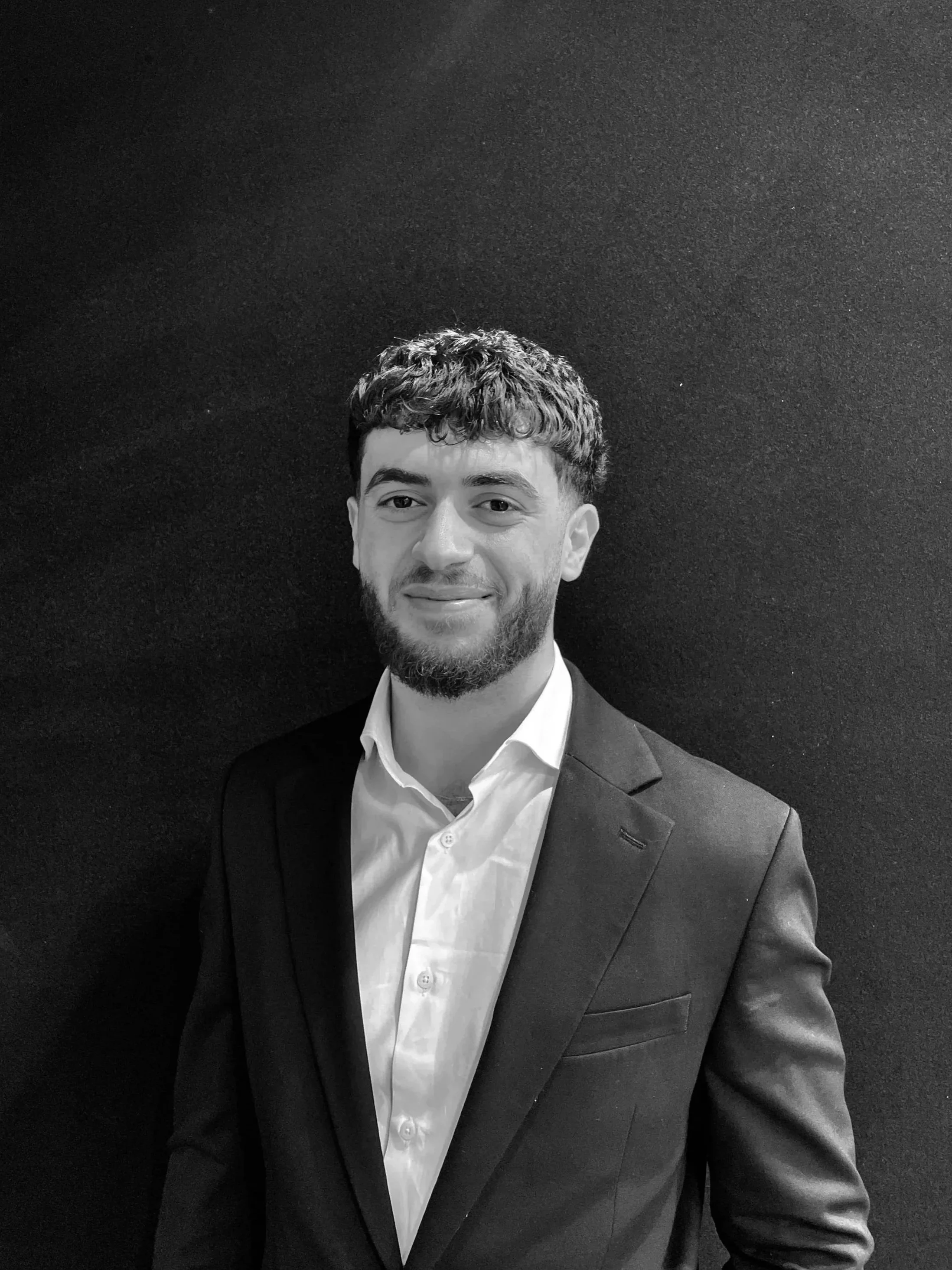 A young man with a beard and short, curly hair, wearing a dark blazer and white shirt, standing against a dark textured background, smiling slightly.