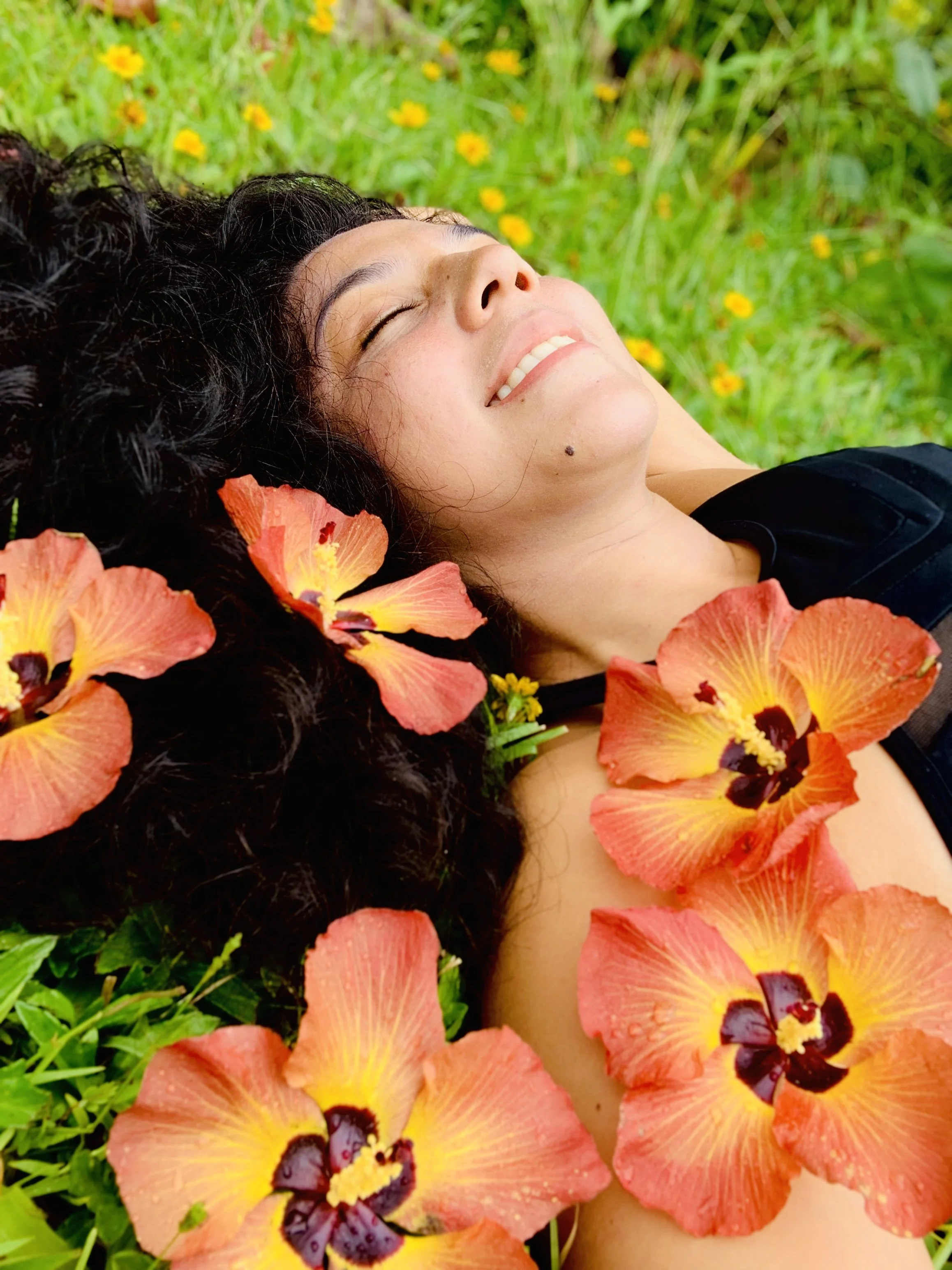A woman with dark curly hair lying on grass surrounded by orange and yellow flowers, smiling with eyes closed.